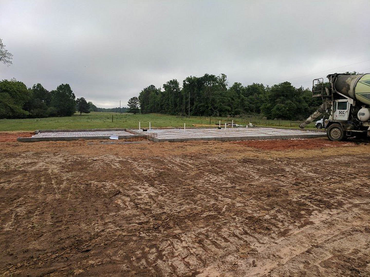 Modern home construction site with crane, concrete mixer truck on dirt field, tire tracks visible in soil, grassy area and trees in background, partially filled pool, close-up of
