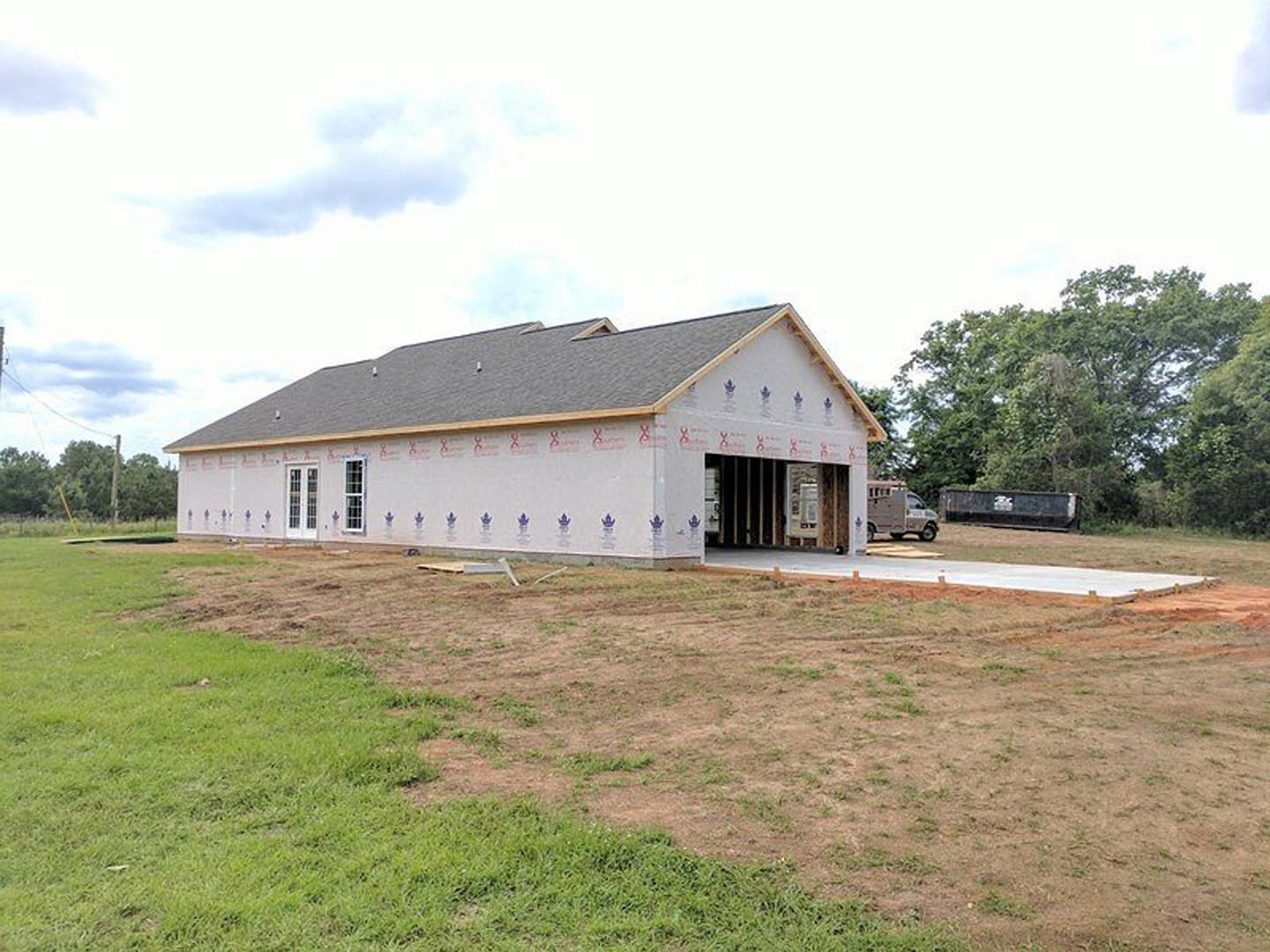 Partially built house with exposed wooden framing, completed shingle roof, construction truck parked on dirt lot, grassy area and trees in background under cloudy sky