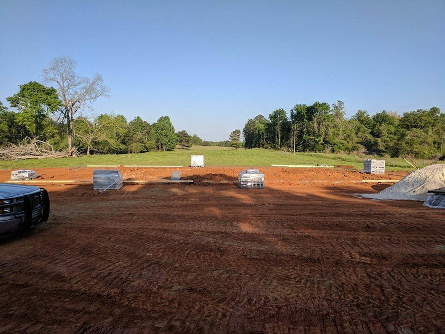 Expansive dirt field bordered by leafless trees, scattered crates, black truck parked in foreground, white square object resting on grass.