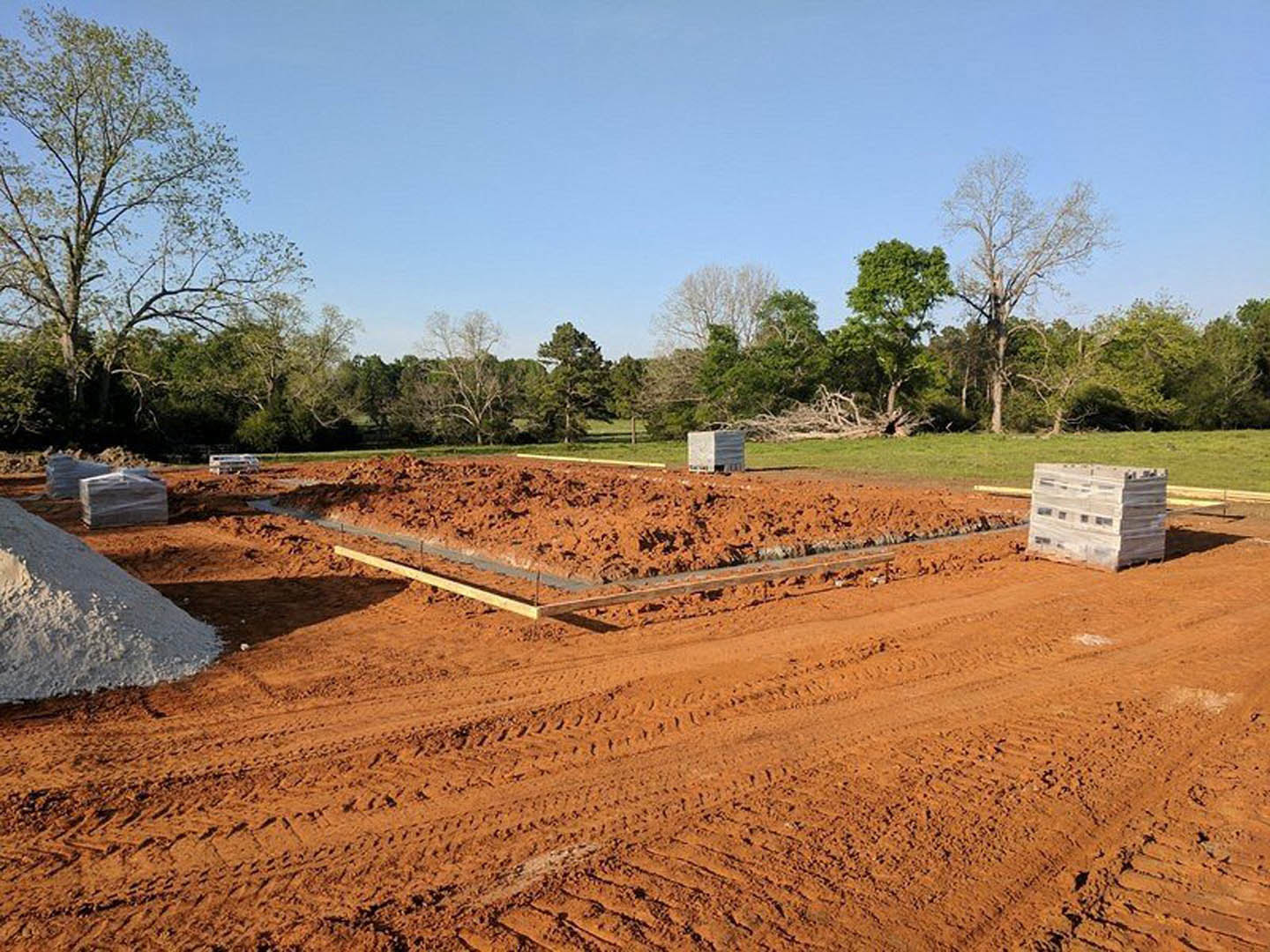 Dirt construction site bordered by trees, stack of pallets and pile of white powder on ground, white box with black stripe, tire tracks visible, blue sky overhead