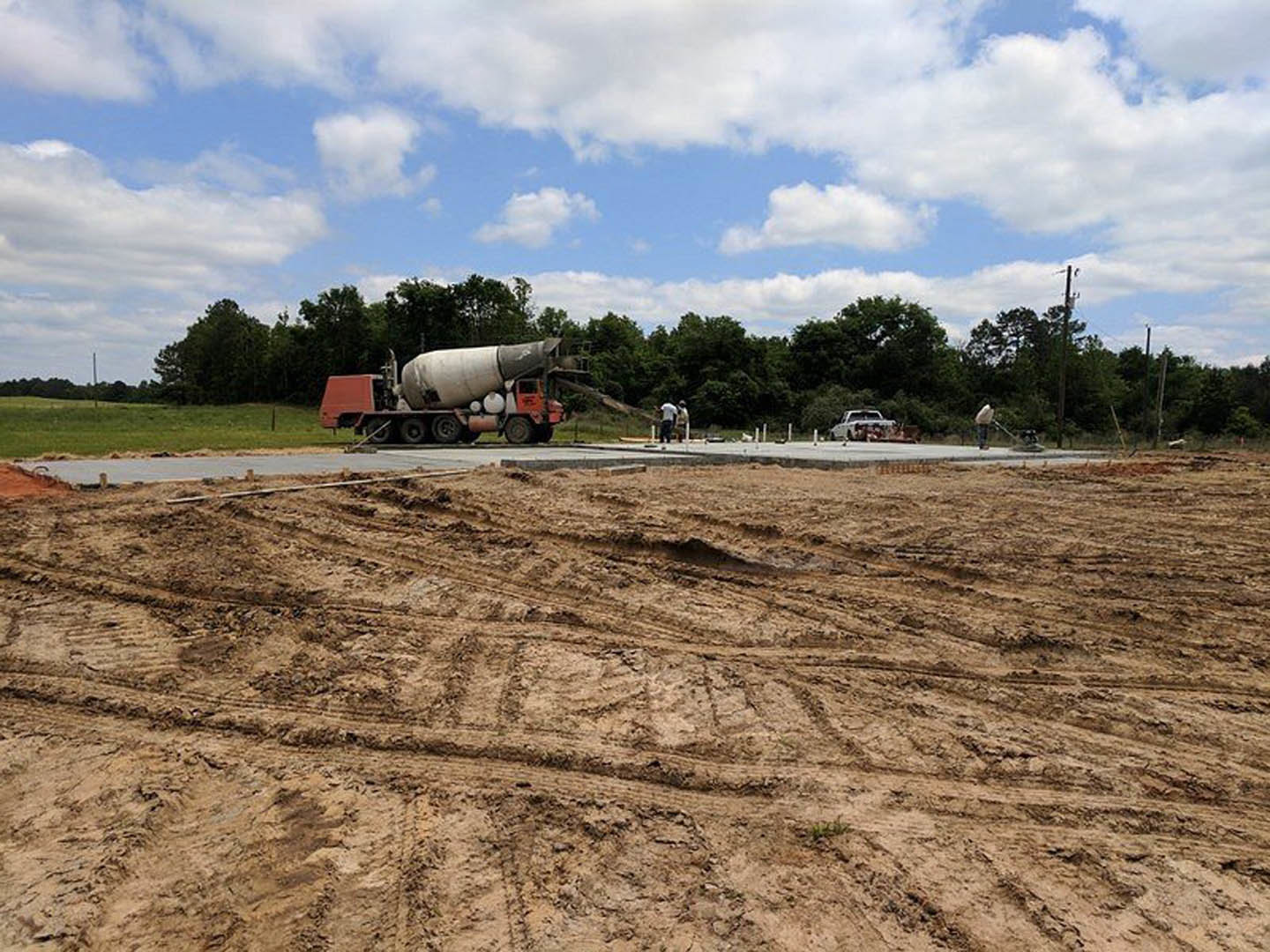 Concrete mixer truck parked on dirt road beside grassy field, tire tracks visible in soil, group of people standing near trees under blue sky with scattered clouds
