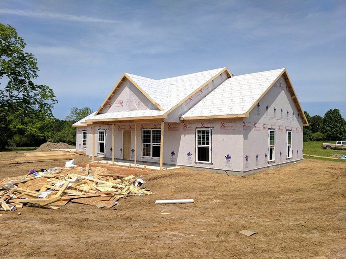 Framed house under construction with exposed insulation, white window frame, and stacked lumber in foreground; leafy tree and historic Robert Frost Farm visible in background