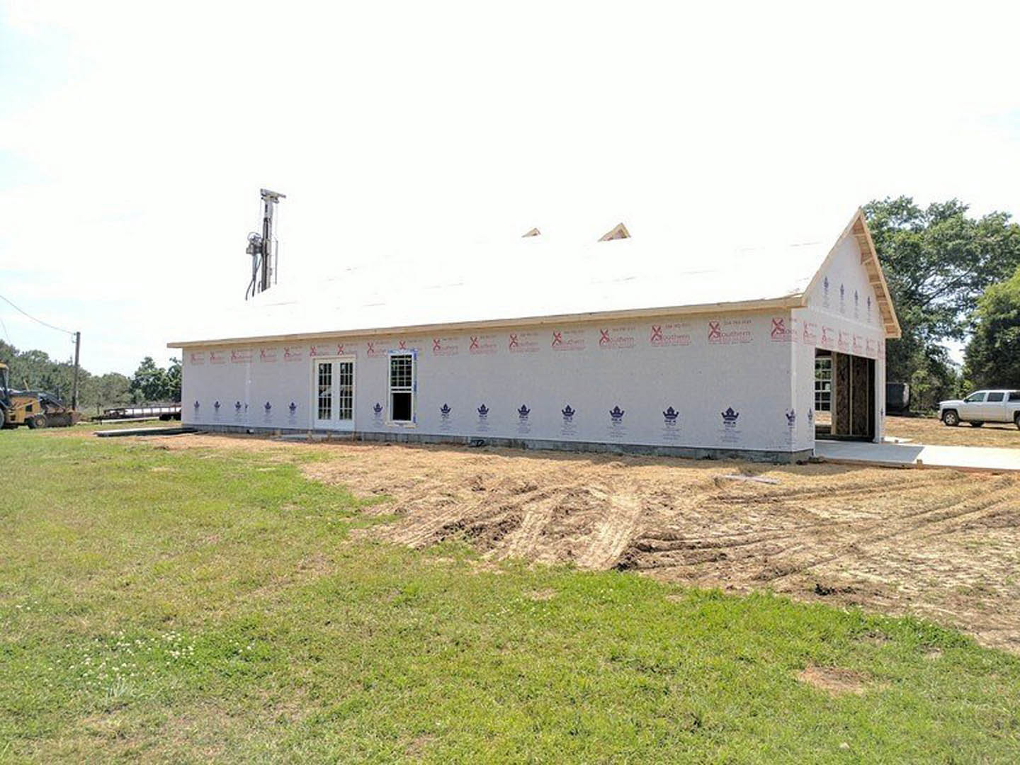 White house under construction with dirt pile in front, grassy field and trees in background, white truck parked nearby, partially visible yellow vehicle, red logo on house