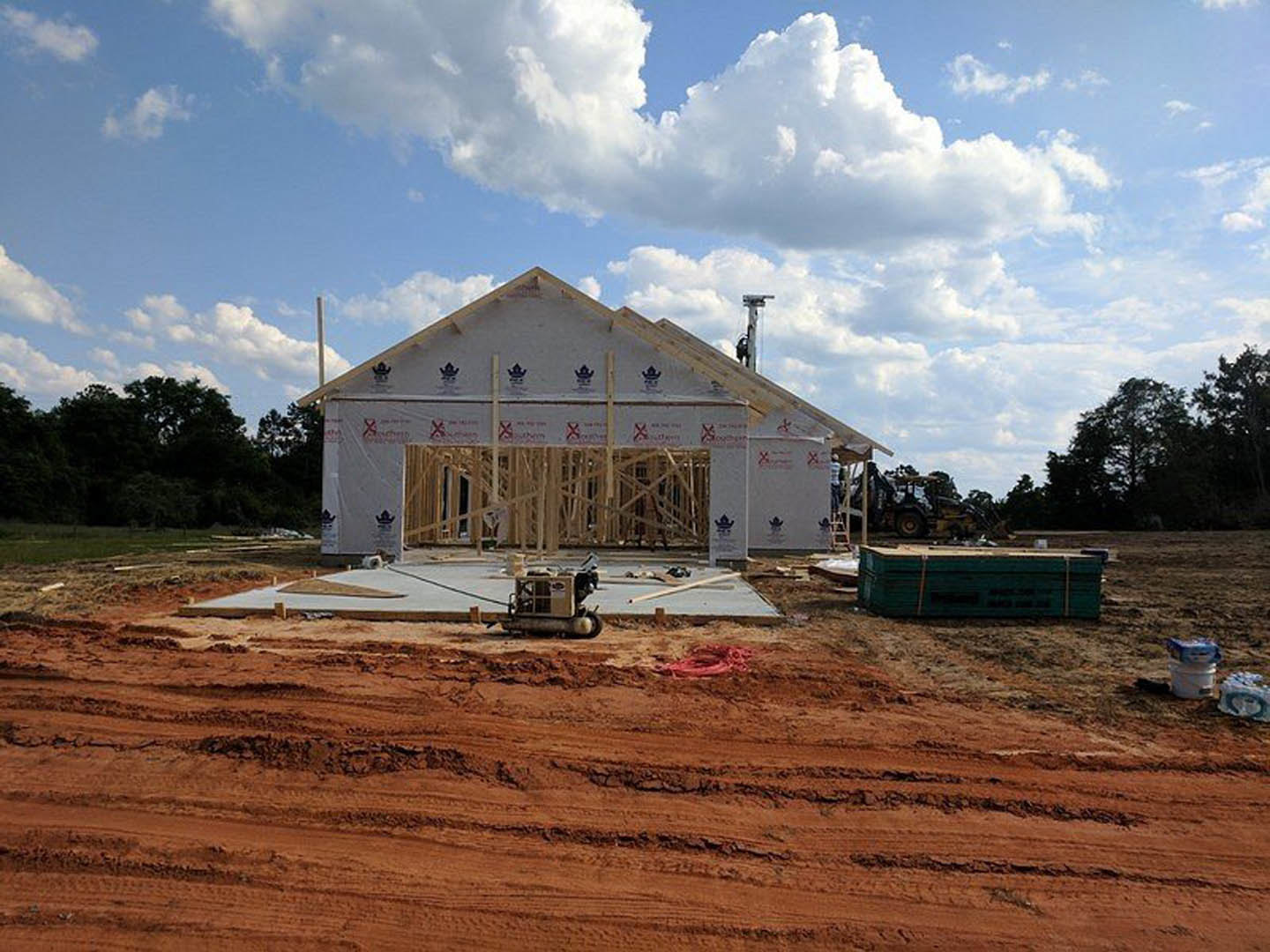 Wood-framed house under construction on a dirt lot, blue sky with scattered clouds overhead, construction equipment and red marking visible on ground
