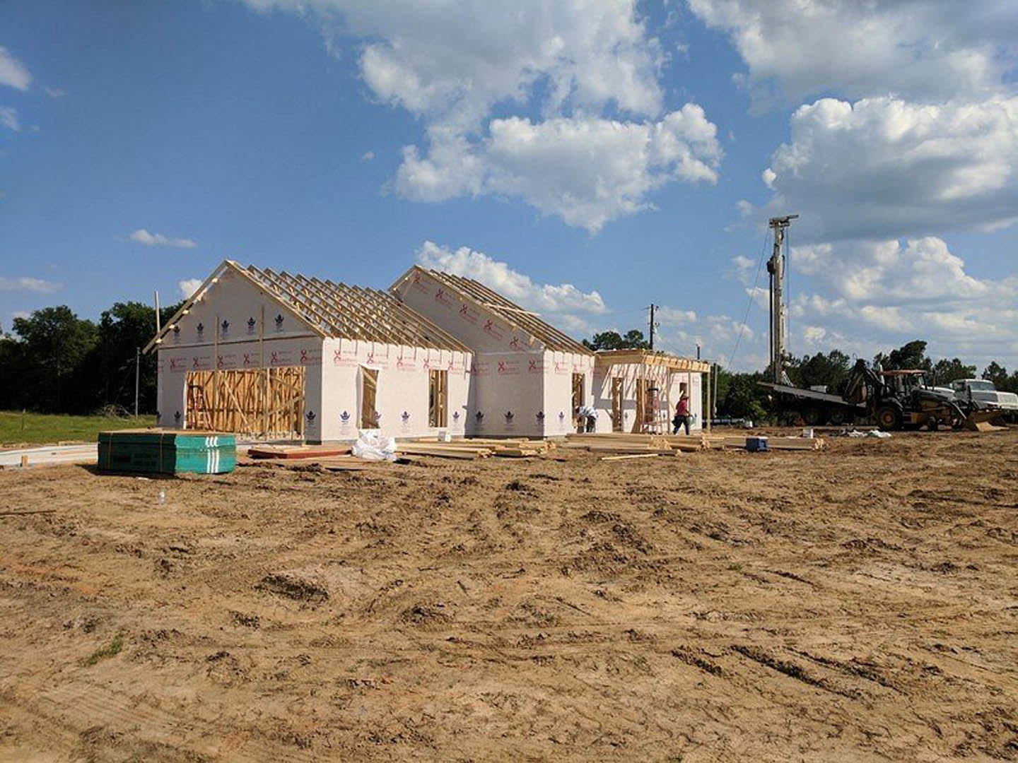 Wood-framed house under construction on a dirt lot with several workers present, surrounded by grass and trees under a cloudy sky