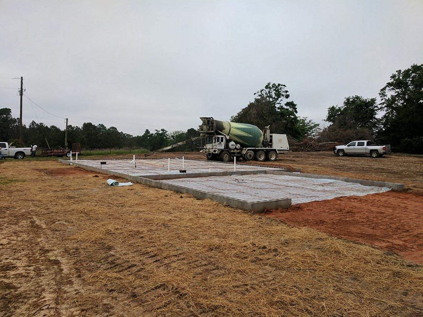 White cement mixer truck parked on dirt lot beside partially built custom home, open truck door and hood visible, surrounded by grass, trees, and construction materials under