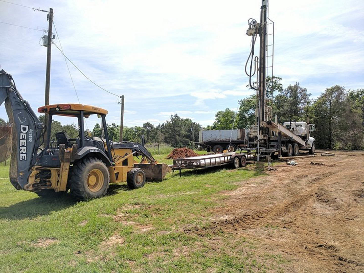 Tractor with backhoe parked beside a pickup truck on grassy lot, telephone poles and trees in background, close-up of tire and truck logo visible