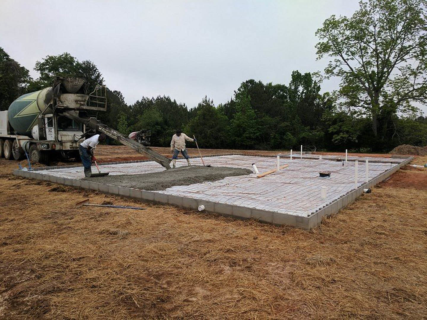 Workers pouring and leveling fresh concrete slab beside a cement mixer truck on grassy lot, surrounded by leafless tree and open sky