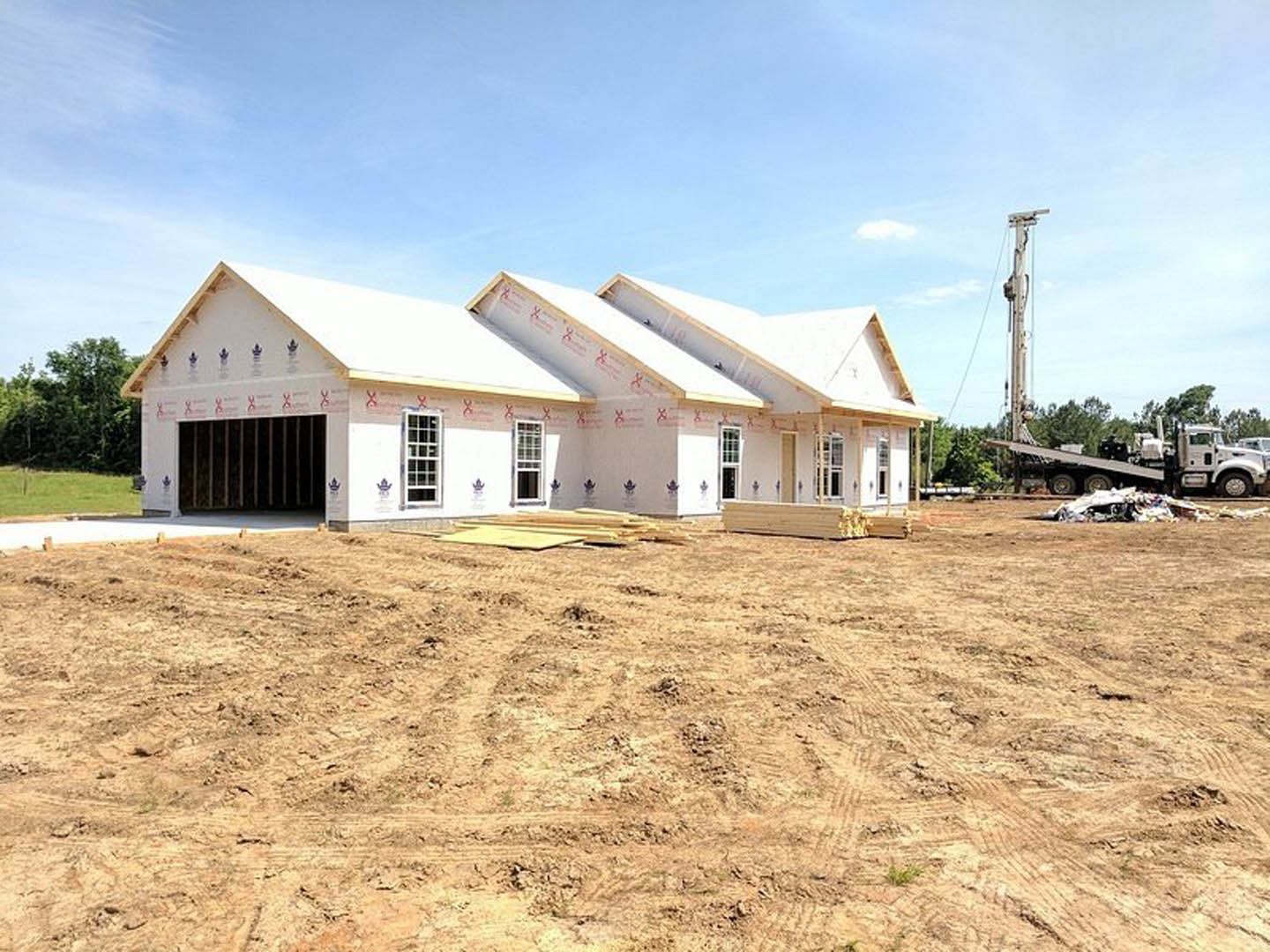 Framed house under construction with exposed wooden beams, shingled roof, surrounded by dirt field, construction materials, and pickup truck; trees and cloudy sky in background