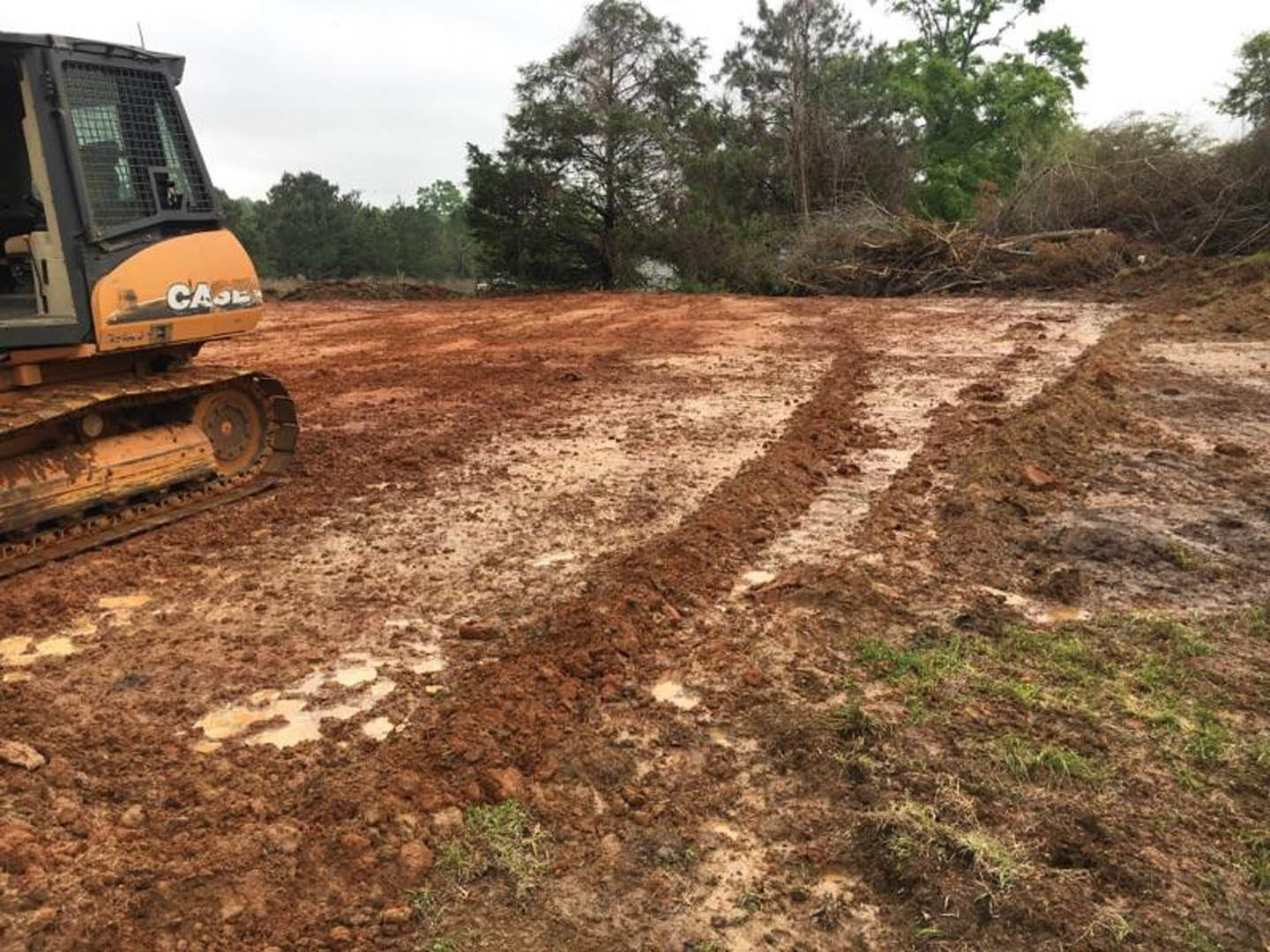 Bulldozer with metal tracks parked on a muddy dirt road, surrounded by grassy field and tall trees under a cloudy sky