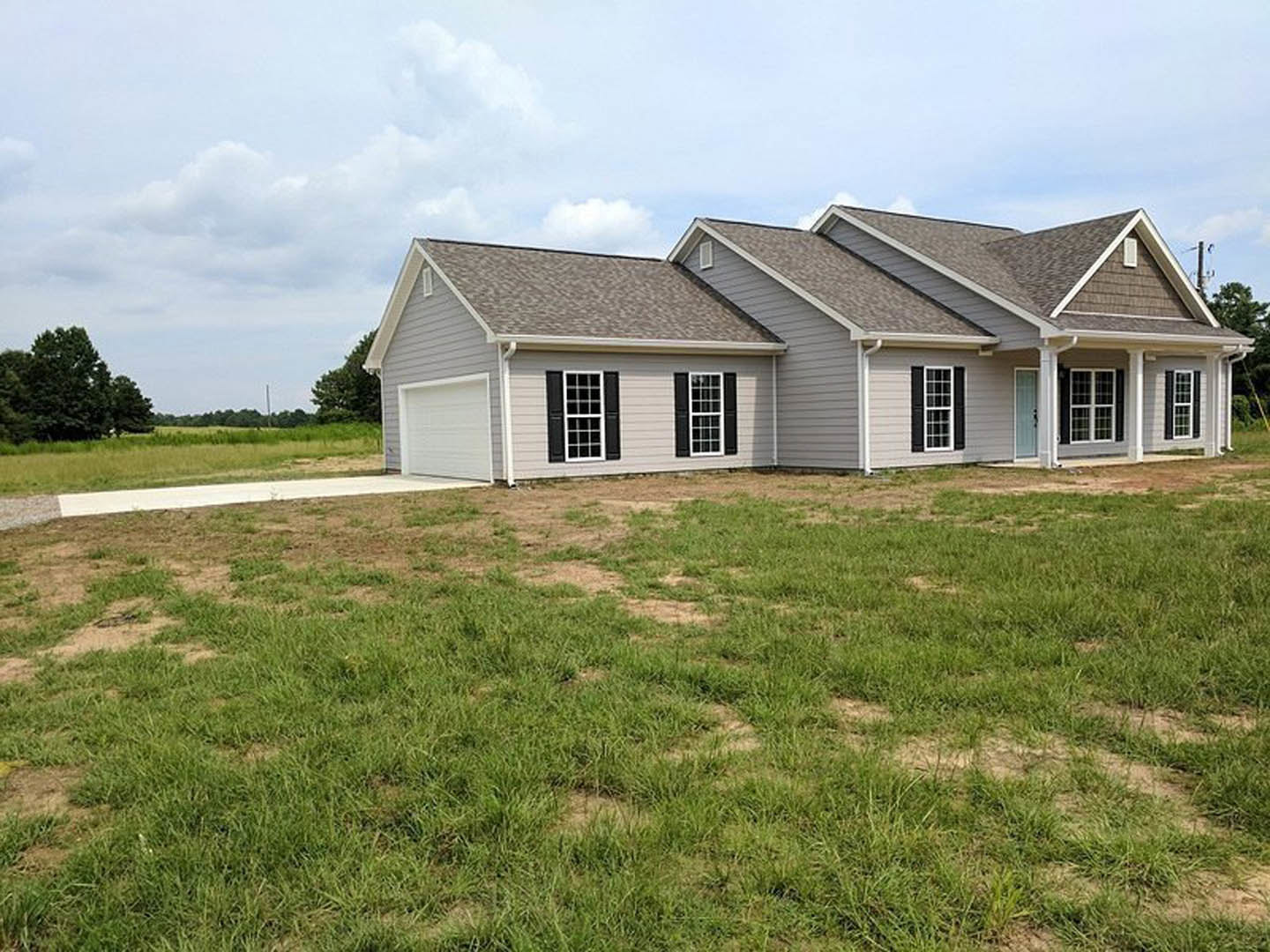 Two-story house with white garage door, gray roof shingles, large windows with white trim, surrounded by green grass lawn and scattered trees under partly cloudy sky