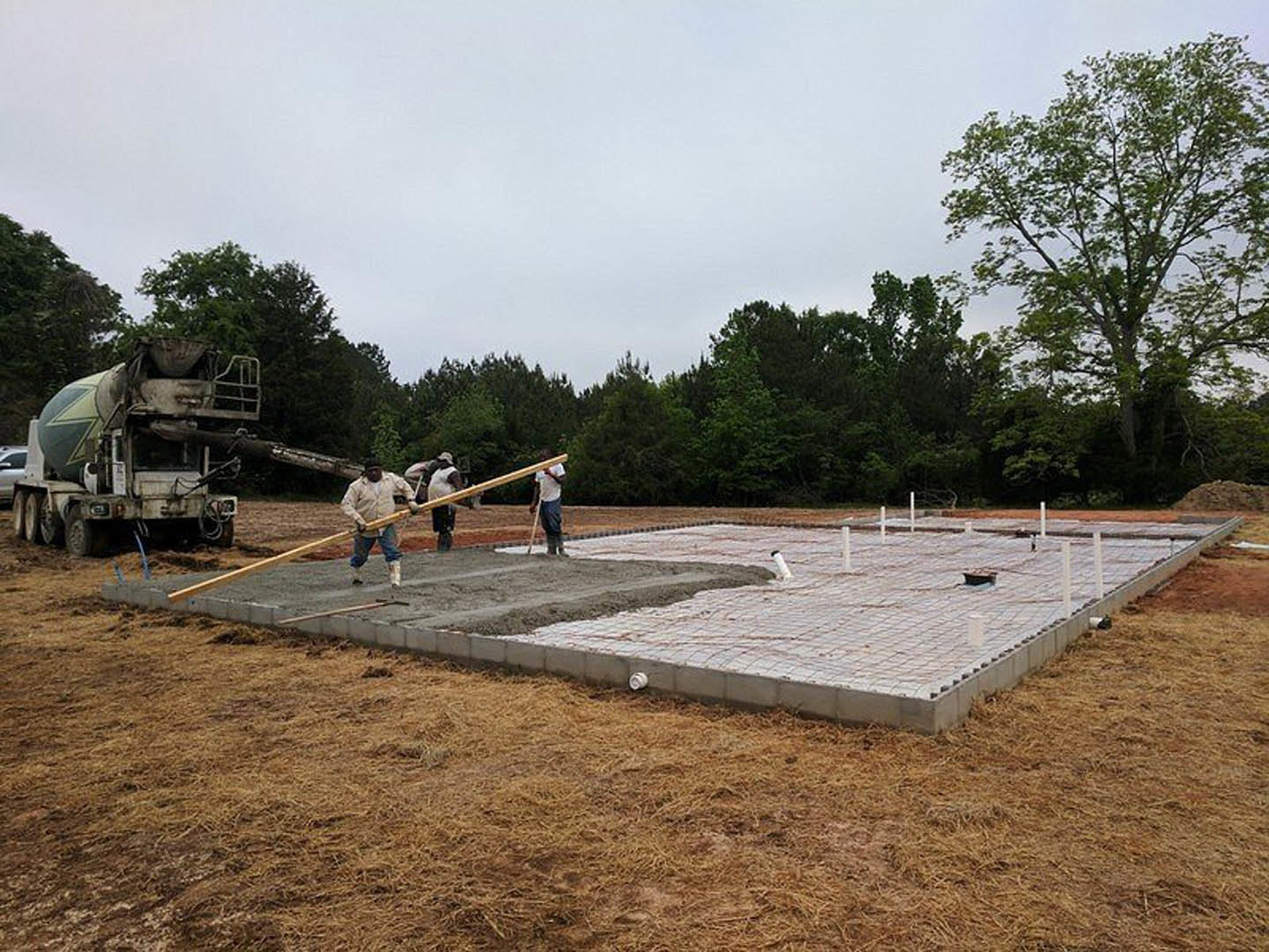 Men pouring and leveling concrete foundation slab with wooden forms, concrete mixer truck parked nearby, leafy tree in background, dirt and grass surrounding construction site