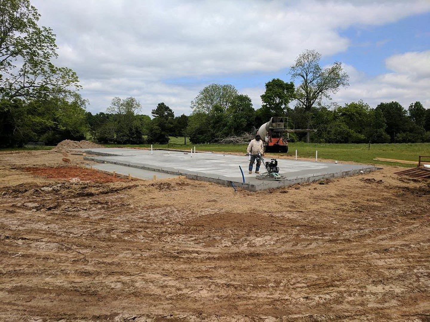 Man in mask and helmet standing beside newly poured concrete slab, surrounded by trees under cloudy sky, tractor parked nearby