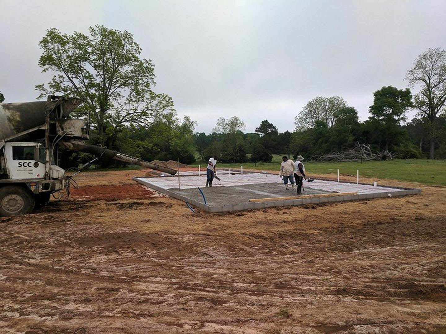 Crew pouring and leveling fresh concrete slab foundation beside parked cement mixer truck, surrounded by dirt, grass, and trees under cloudy sky