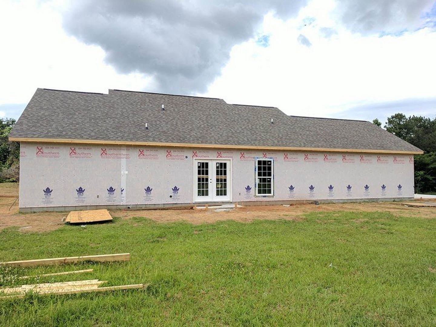 Partially built farmhouse with white double doors, glass windows, exposed wood framing, and green grass field in foreground under cloudy sky