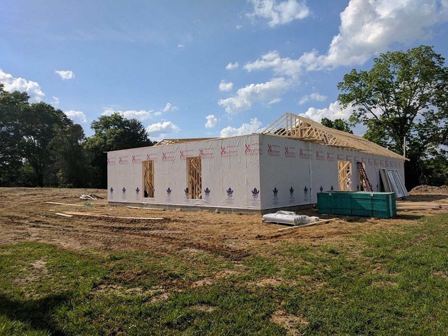 Framed house under construction with white walls covered in plastic, green wall section, large window, surrounded by grass and trees beneath a blue sky with clouds