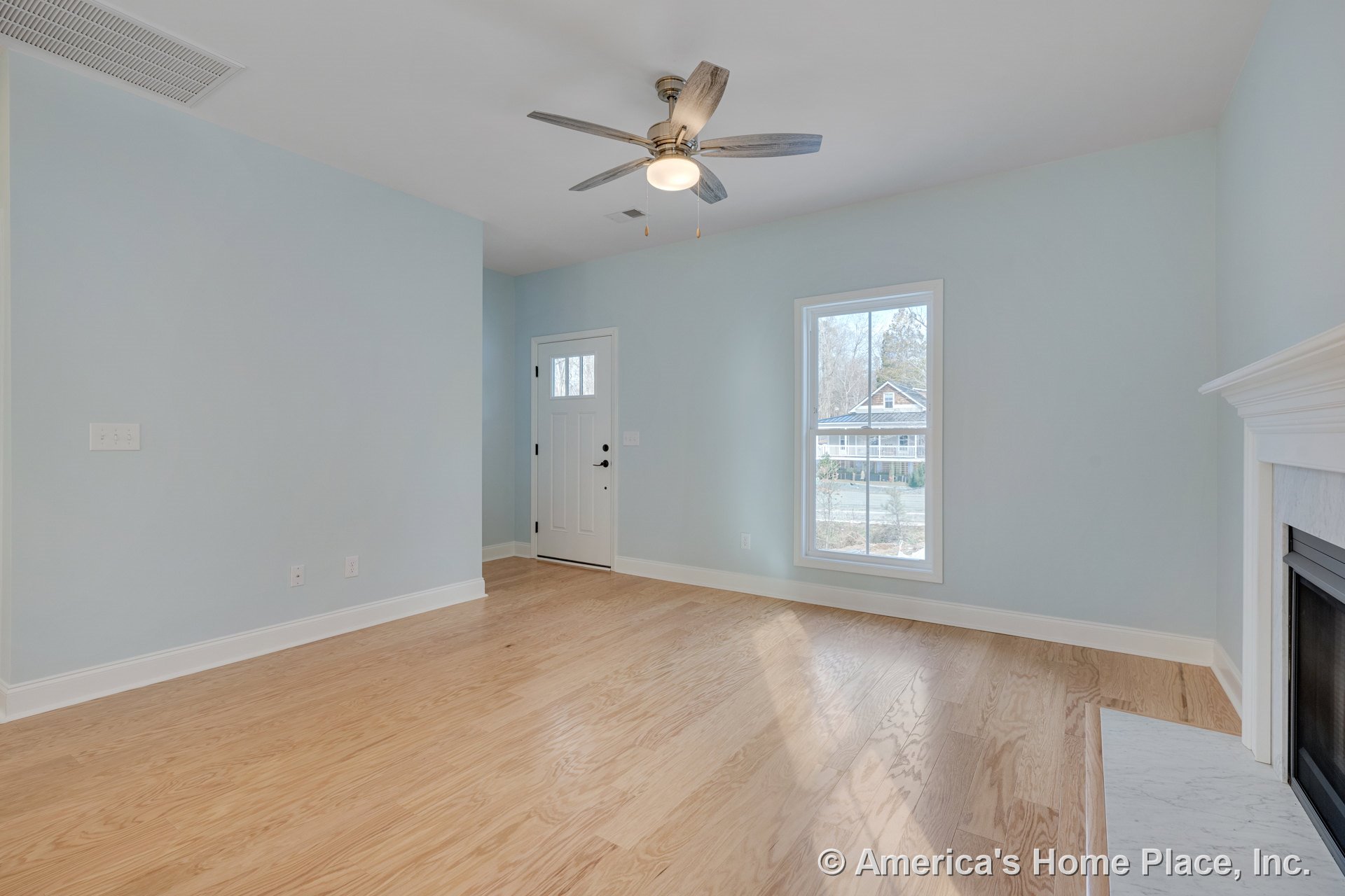 Light wood flooring and white baseboards, single large window, white front door with glass insert, ceiling fan with built-in lighting, white fireplace with marble surround, light