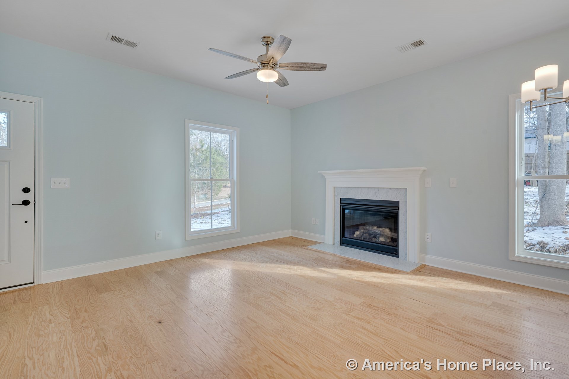 Gas fireplace with white mantel centered on painted wall, light wood flooring throughout, large windows with white trim, ceiling fan with light fixture, built-in chandelier, entry