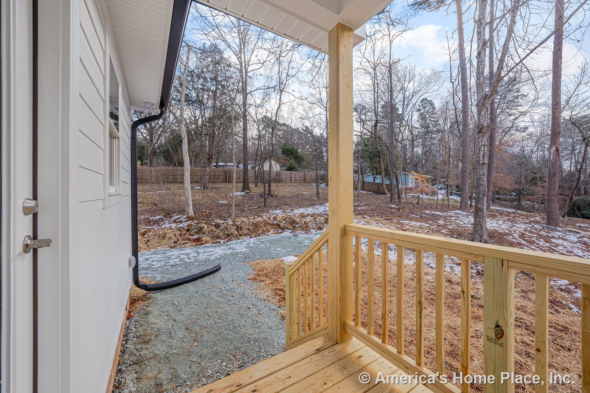 Small covered porch featuring unfinished wood railing and decking, exterior siding, trim, and door hardware, overlooking a tree-lined backyard.
