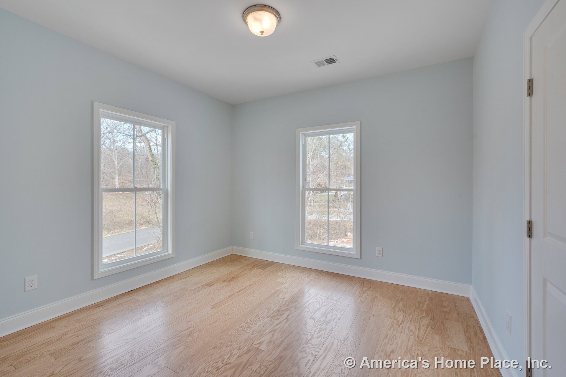 Bedroom with light wood flooring, two large double-hung windows, white baseboards, flush mount ceiling light, light blue painted walls, and white door and window trim.