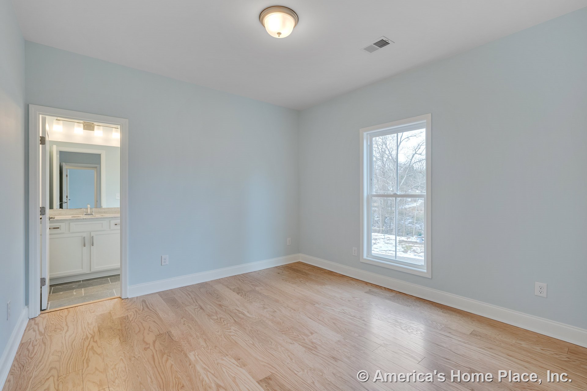 Bedroom with light wood flooring, single window framed in white trim, flush mount ceiling light, and white baseboards; en suite bathroom visible through doorway features white