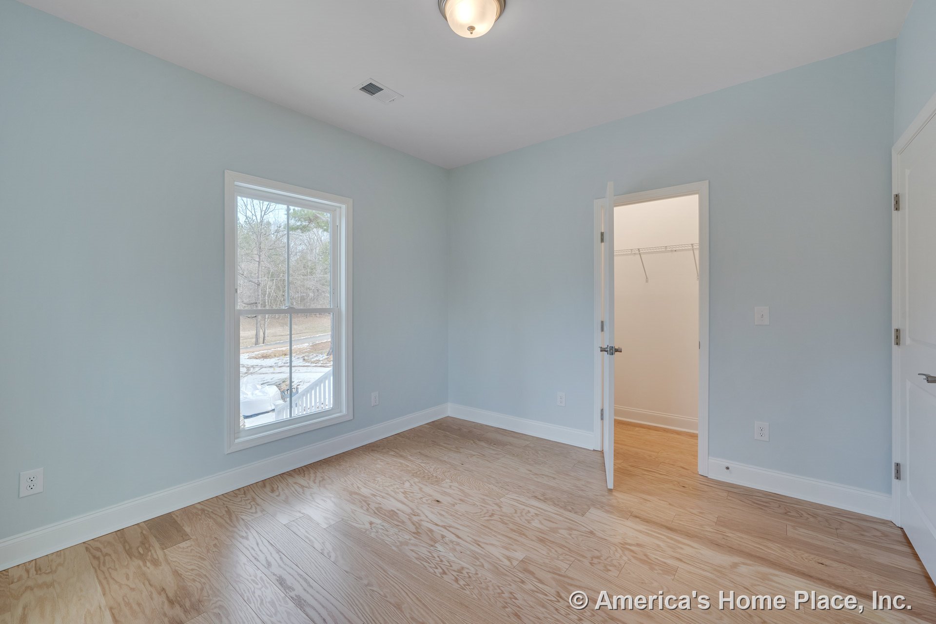 Bedroom with light wood plank flooring, pale blue painted walls, large window with white trim, flush mount ceiling light, white baseboards, and walk-in closet featuring wire