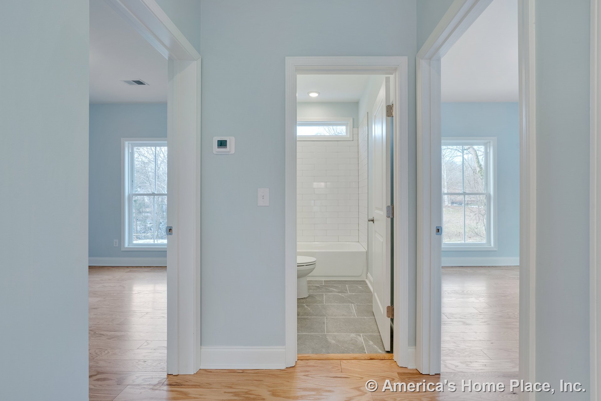 Hallway with wood flooring and wall-mounted thermostat leads to bathroom featuring white subway tile surround and gray floor tile, flanked by two bedrooms with large double-hung