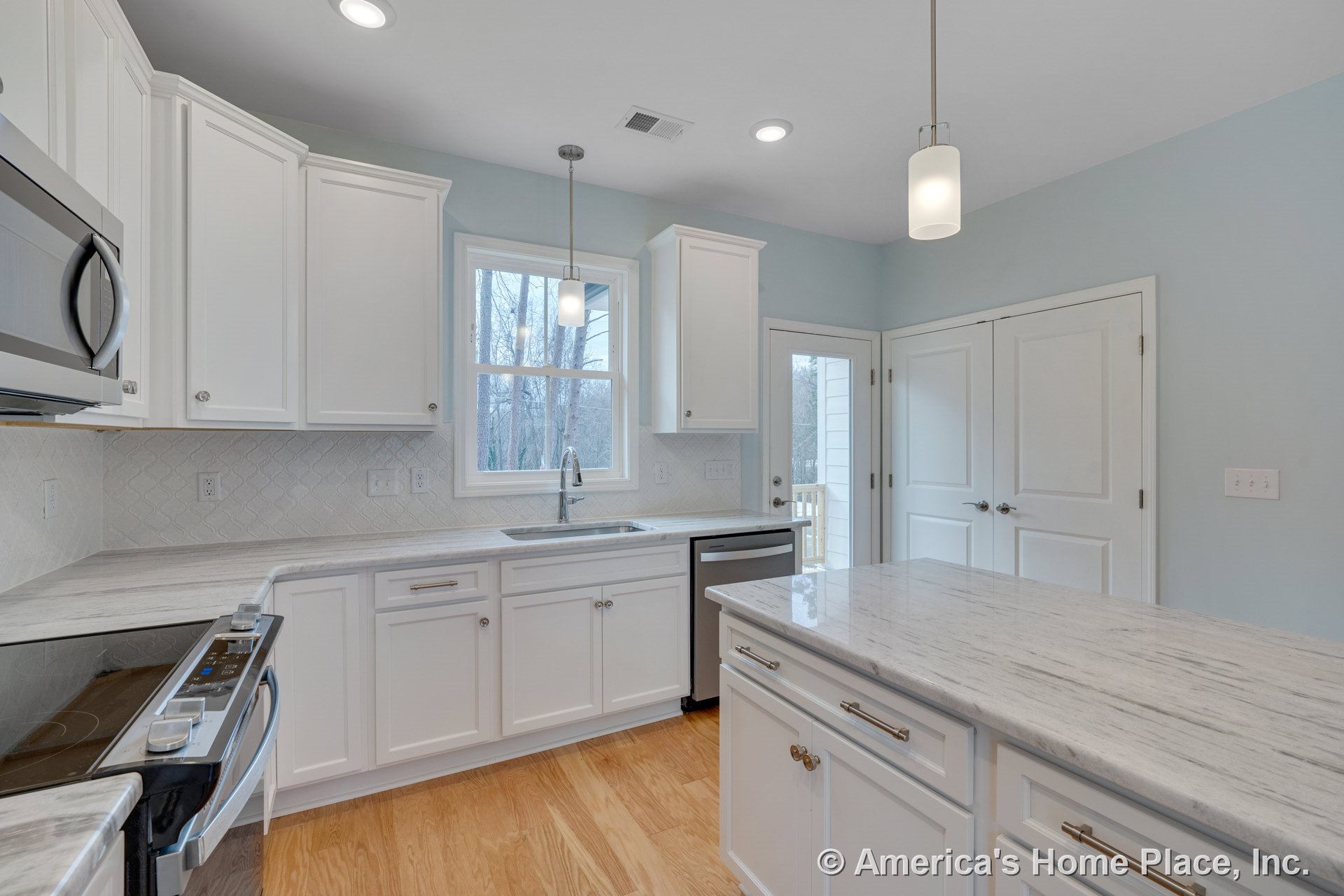 White upper and lower cabinets with marble island countertop, tile backsplash, stainless steel built-in appliances, pendant and recessed lighting, wood flooring, window above sink