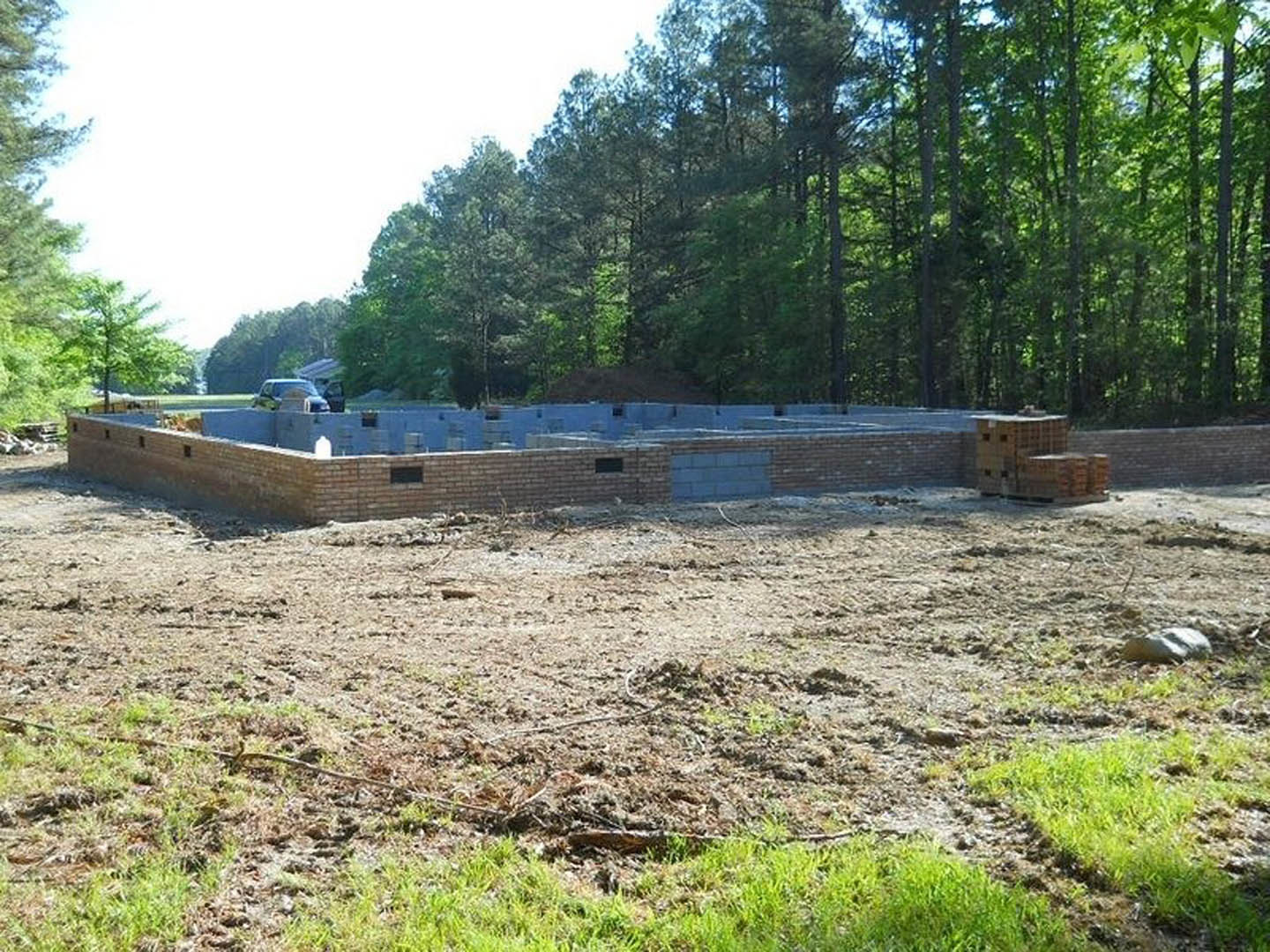 Partially built brick home with exposed blue and black masonry, dirt yard scattered with grass and construction materials, wooded area in background