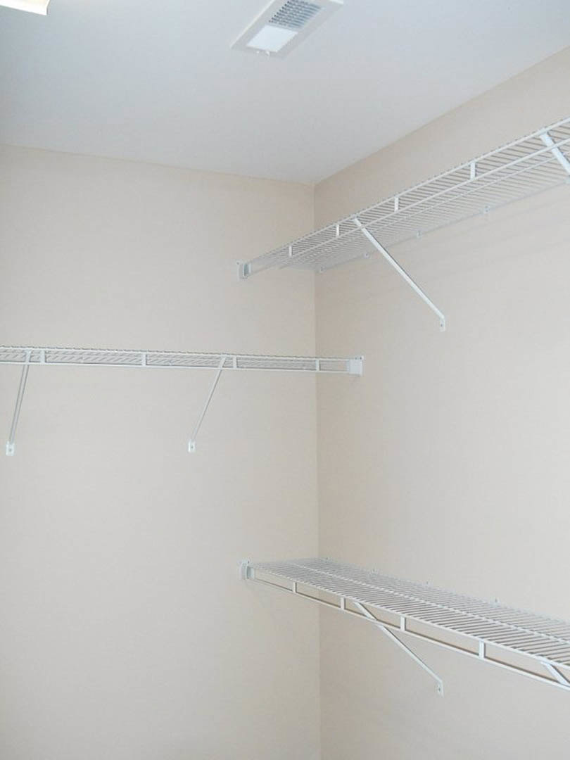 White built-in shelving unit against a plaster wall in a closet room, with a ceiling vent and ceiling fan visible above.