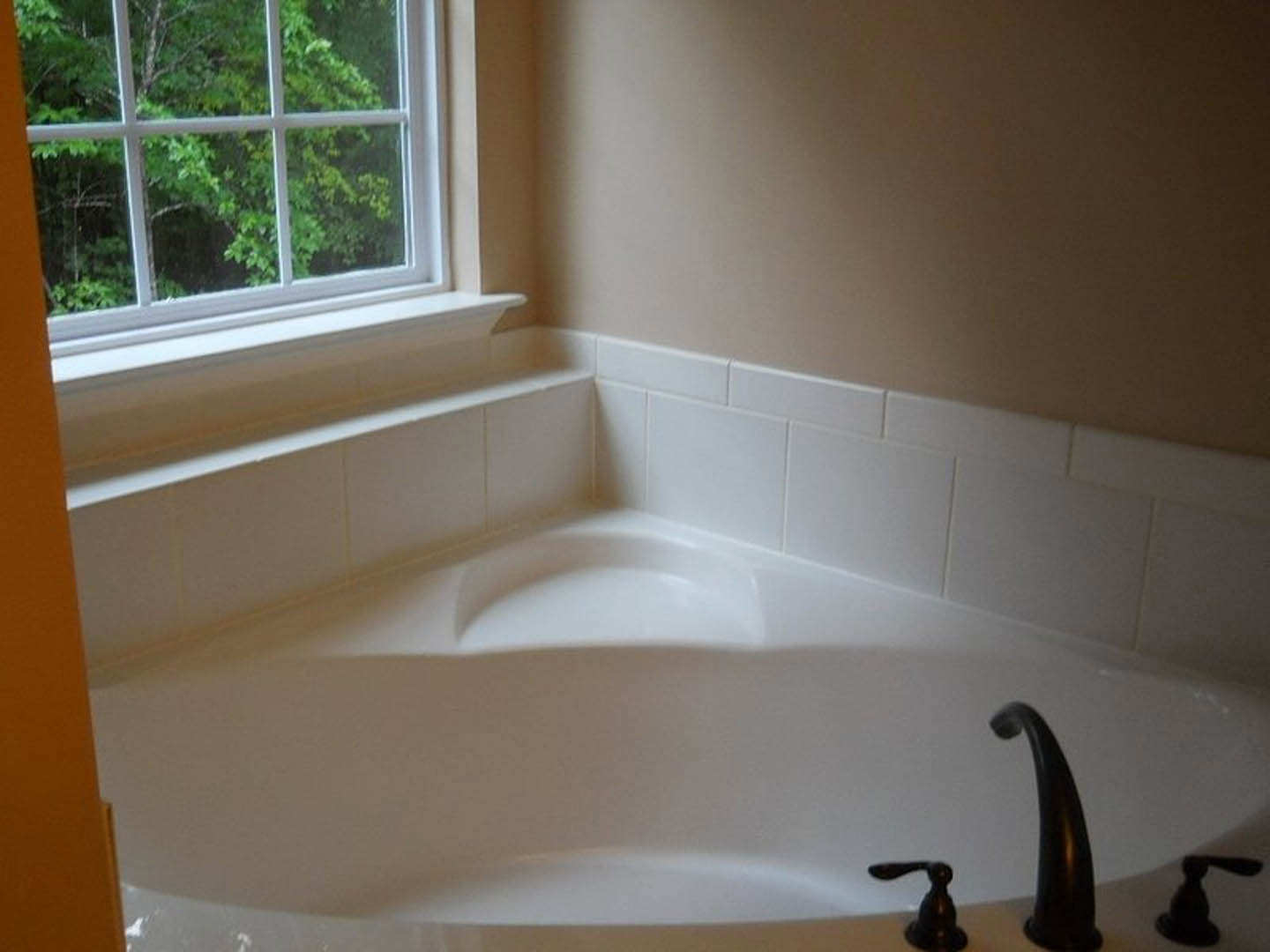 White freestanding bathtub with black handle in bathroom corner, white tile wall, tan painted wall, multi-pane window, chrome faucet