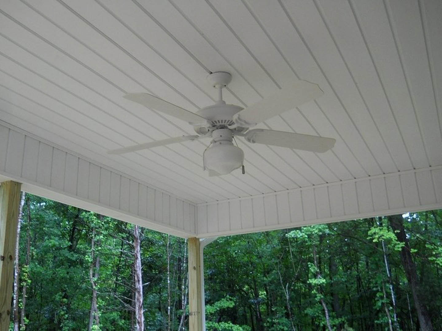 White ceiling fan with integrated light fixture mounted on a covered porch, white pillars and trim, leafy trees visible in the background through windows.