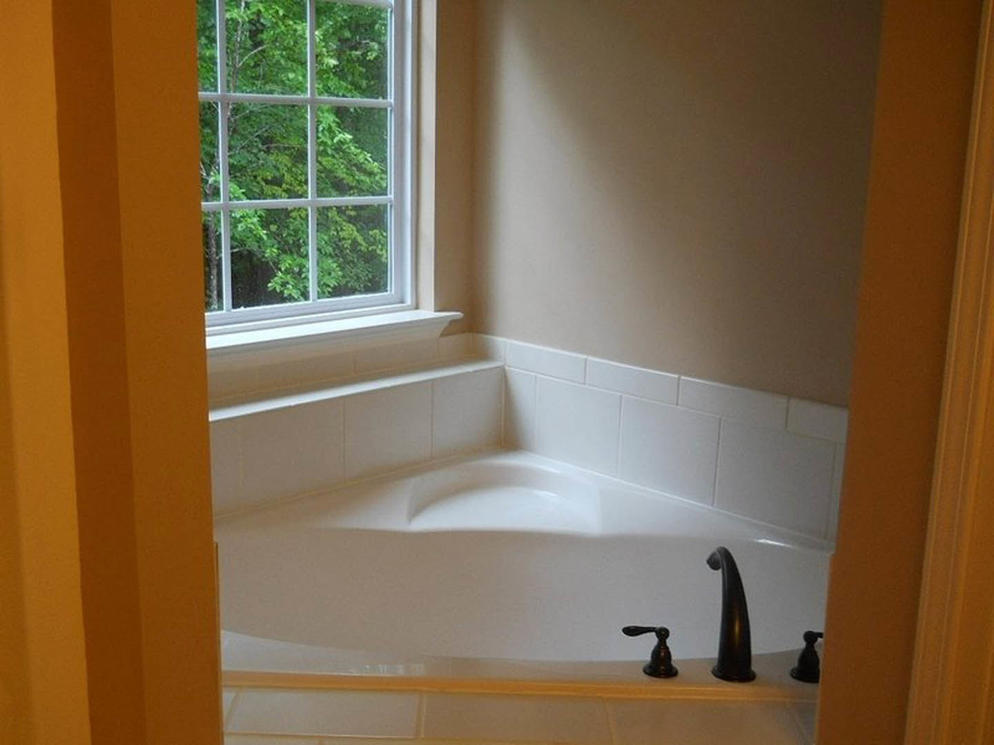White freestanding bathtub with black handle positioned in a bathroom corner, large window above offering view of green trees, chrome faucet, light-colored walls