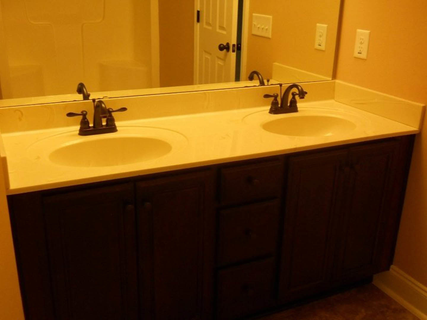 Bathroom with two undermount sinks set in a quartz countertop, large frameless mirror above, chrome faucets, white cabinetry, tile flooring, visible wall outlet and white door.