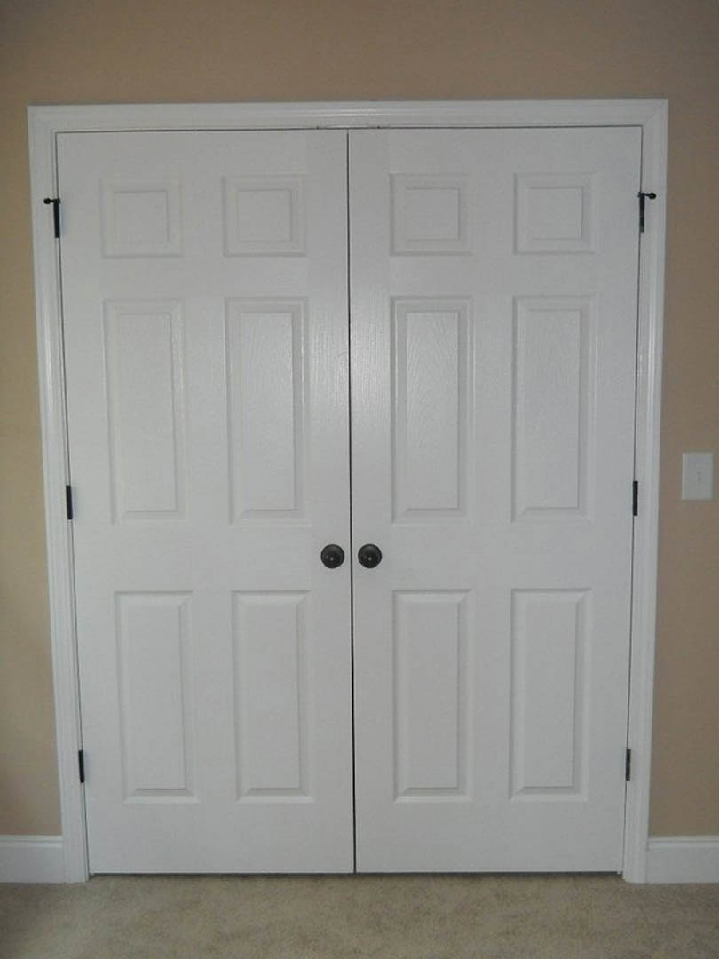 White double doors with black handles set in a smooth white wall, light-colored flooring visible at the base, simple modern interior detailing