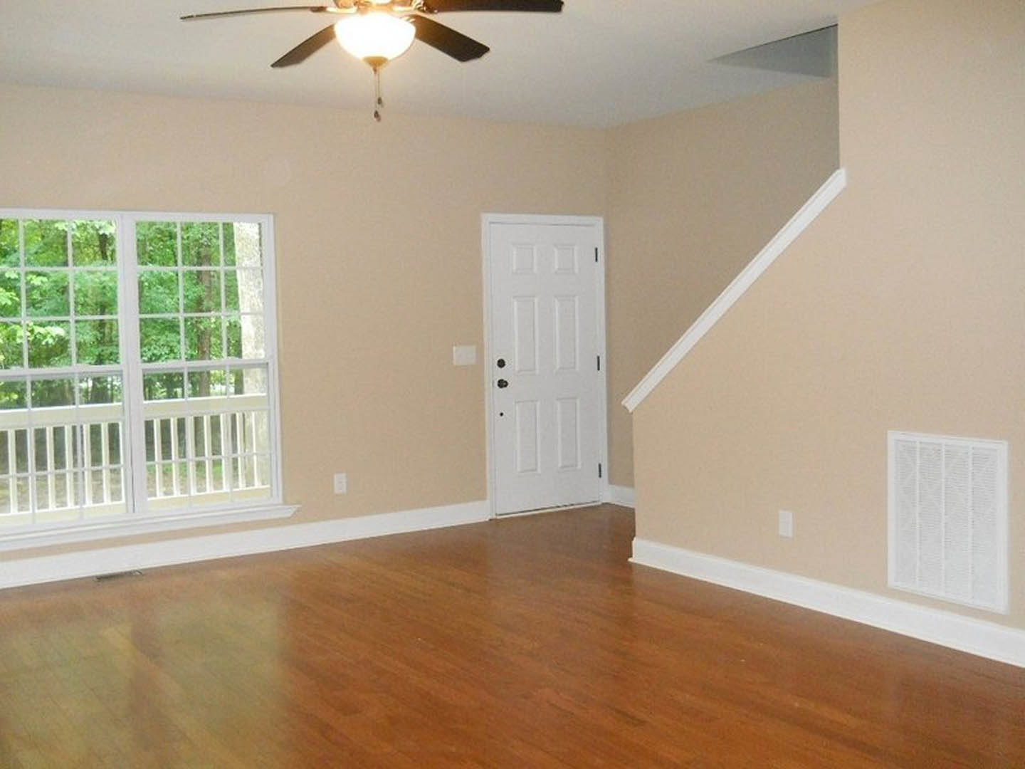 Hardwood floor with white baseboard trim, white door with black hardware, ceiling fan with dark blades, window showing leafy trees, white air filter on wall