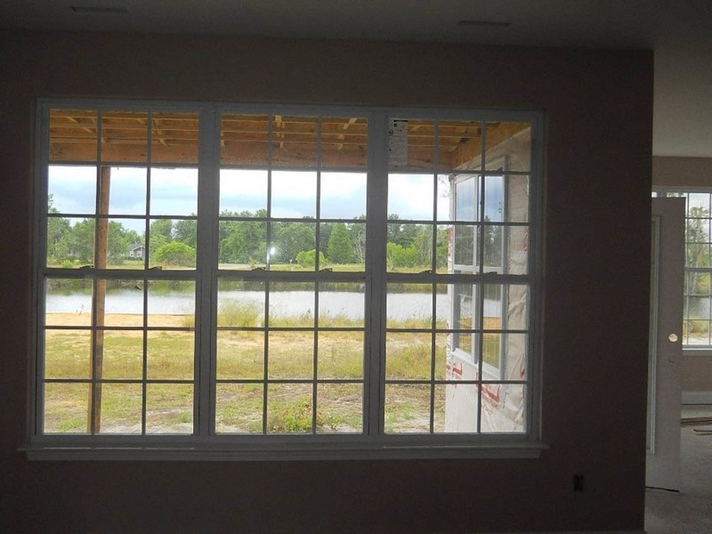 Large multi-pane window framed by white trim, overlooking a serene lake and neighboring house, set in a room with light walls and hardwood flooring.