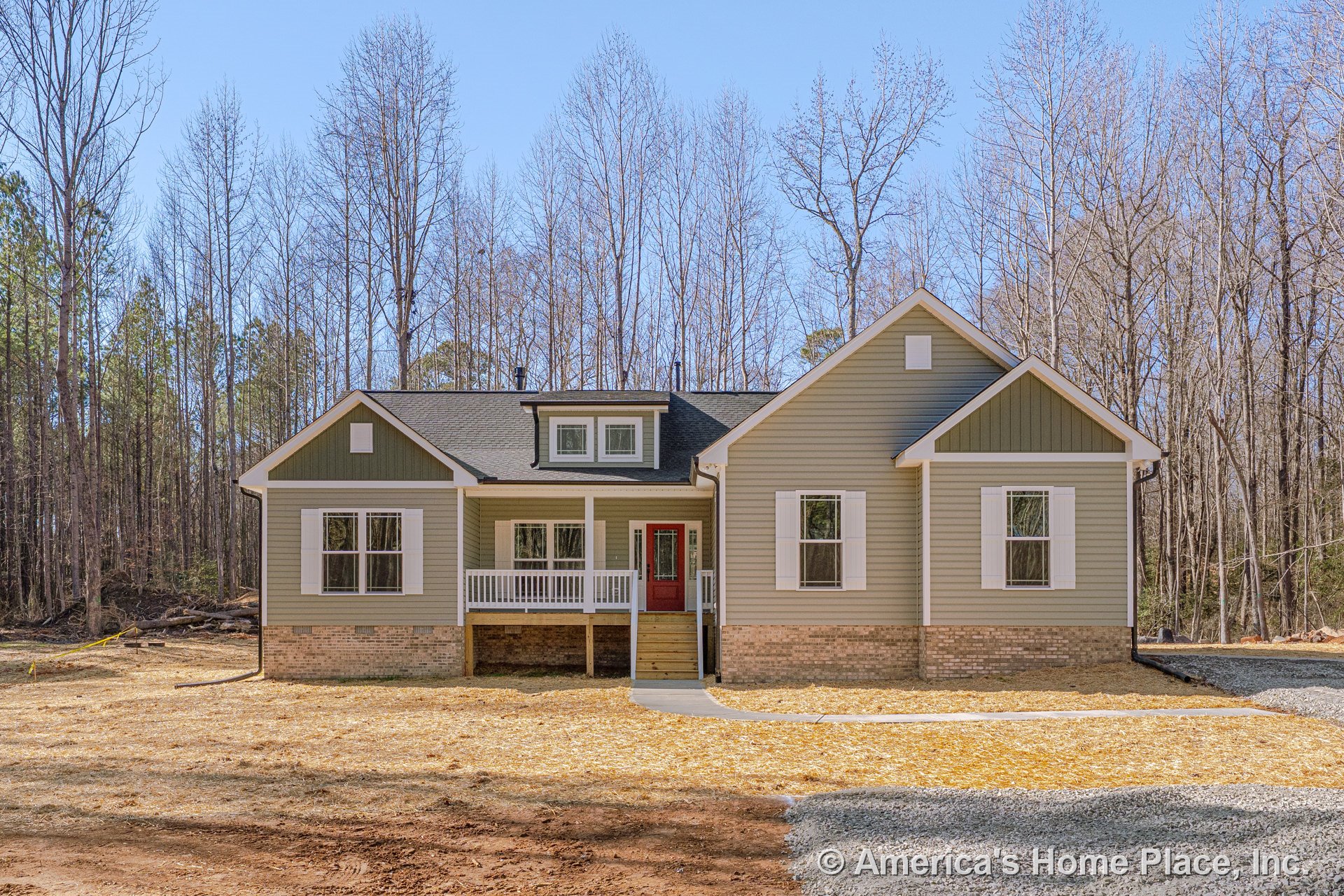 Covered front porch with white railings and red entry door, green horizontal siding, white window trim and shutters, brick foundation, gable roof with dormer, and concrete walkway.