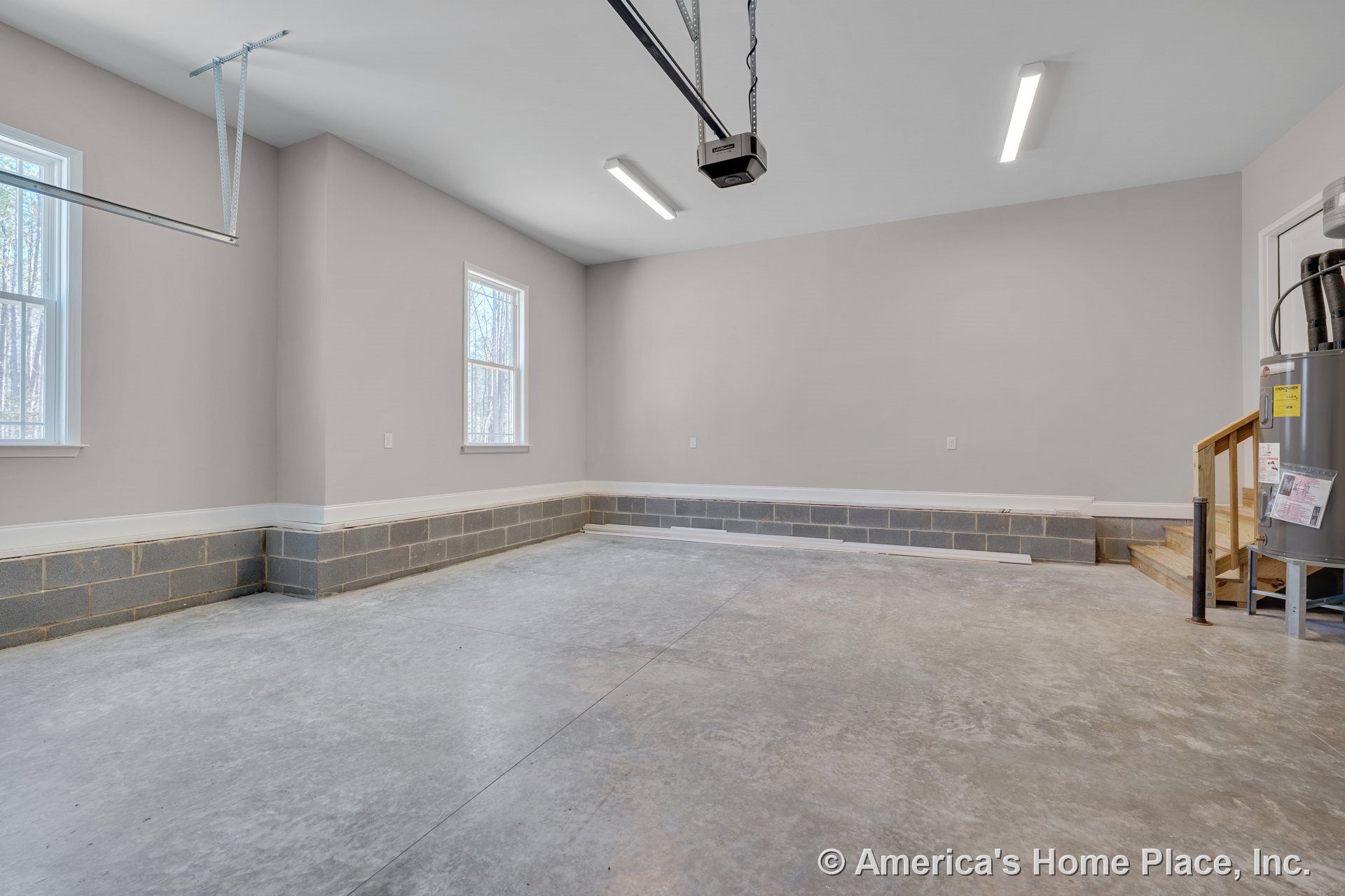Garage interior with concrete flooring, cinder block foundation walls, two windows providing natural light, fluorescent ceiling lights, garage door opener, and wooden steps leading