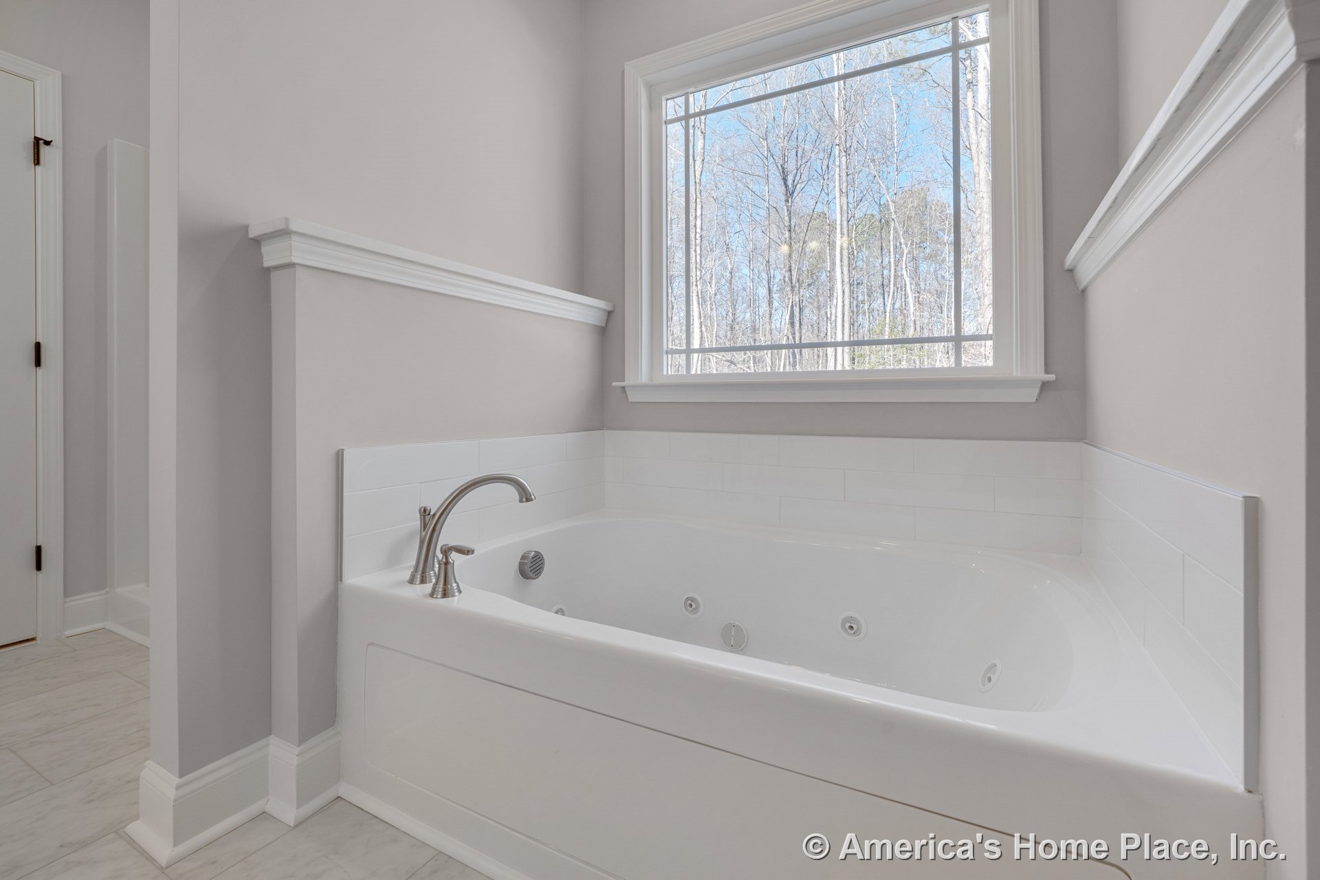 Built-in jetted soaking tub with chrome faucet set beneath a large window, surrounded by white tile backsplash and marble-look floor tile; neutral wall paint and white window trim