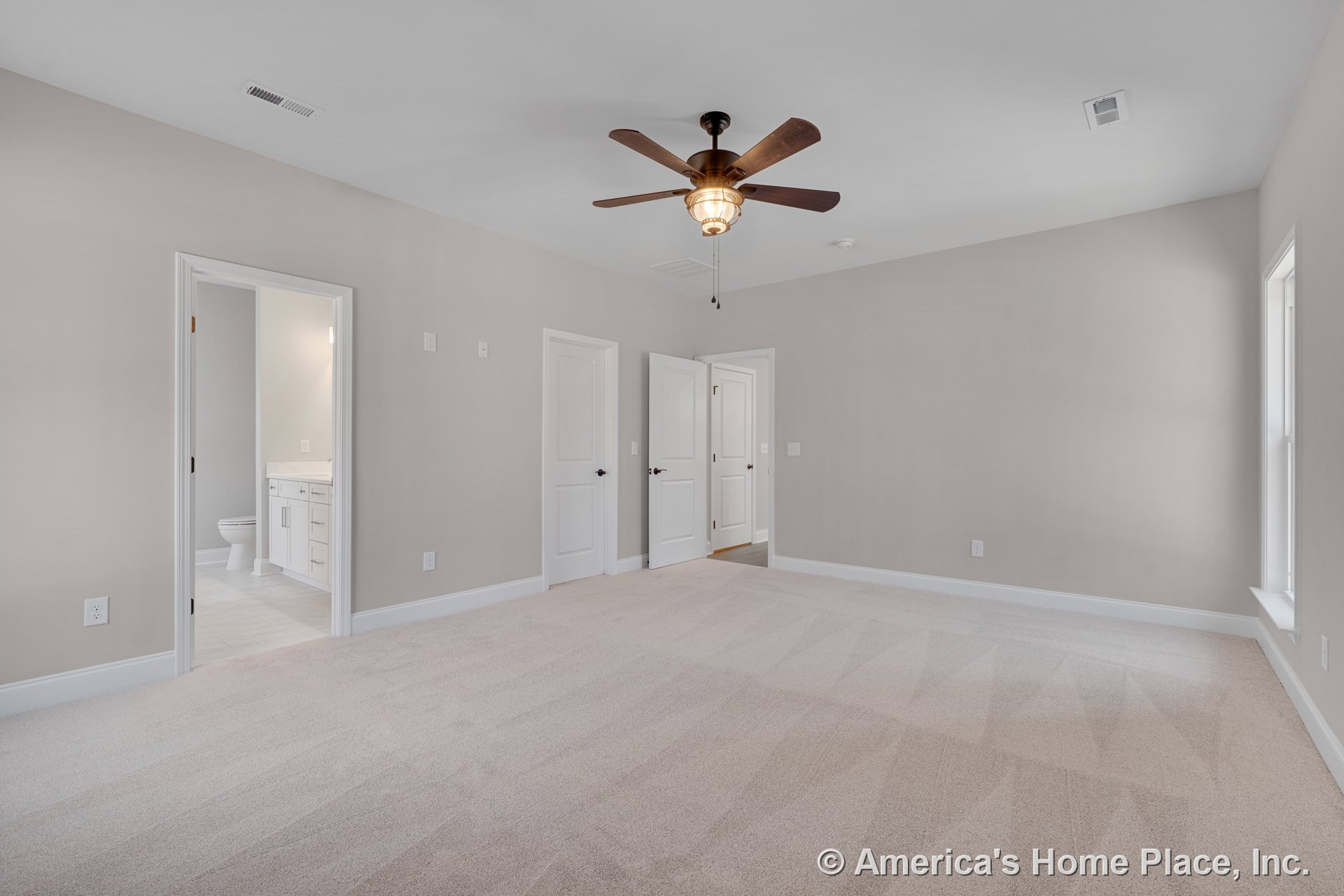 Spacious bedroom with neutral carpet flooring, ceiling fan and built-in light, large window, white trim and baseboards, multiple paneled doors, and en suite bathroom featuring