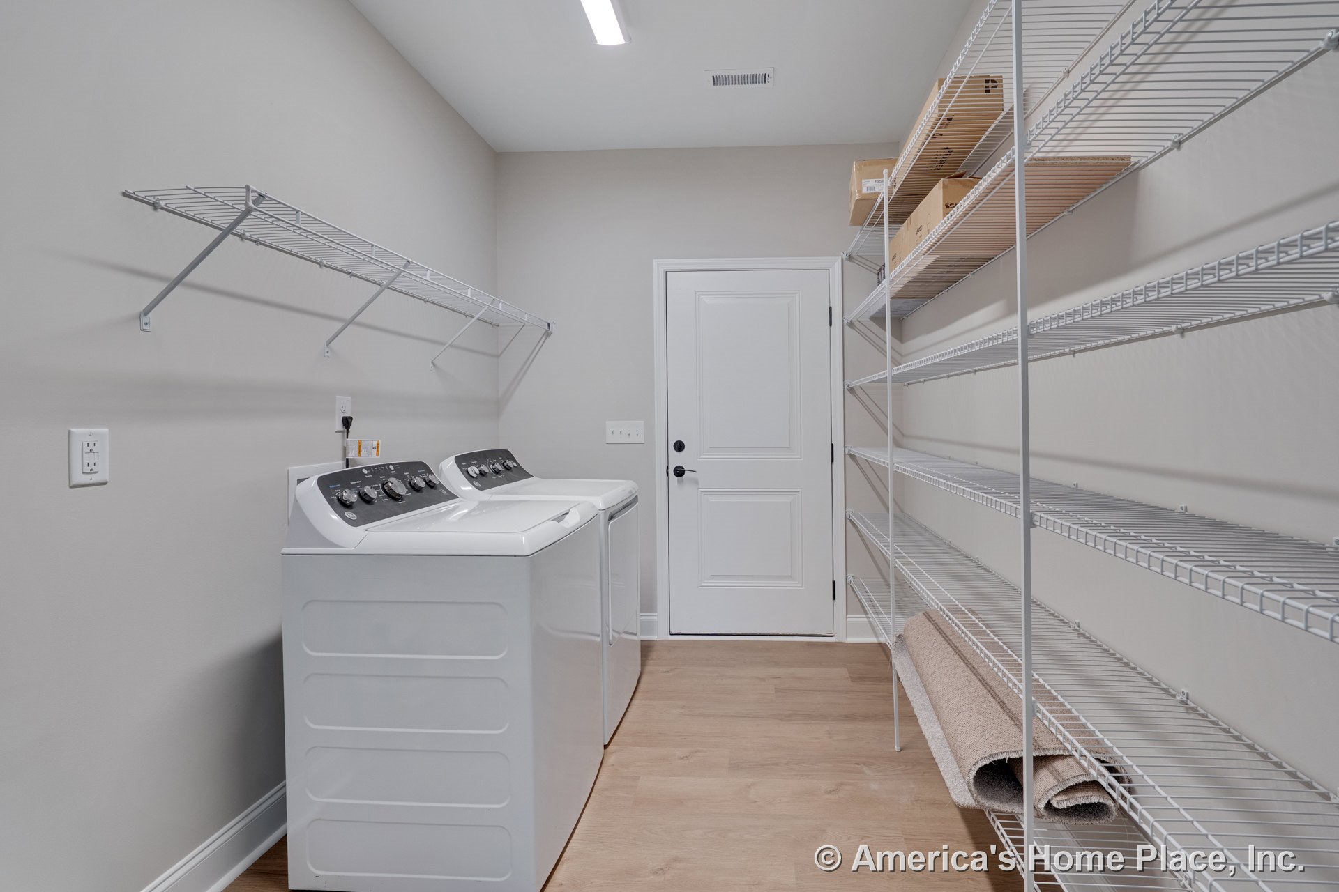 Laundry room with wall-mounted wire shelving above a side-by-side full-size washer and dryer, light wood flooring, recessed ceiling light, white paneled door, and neutral painted