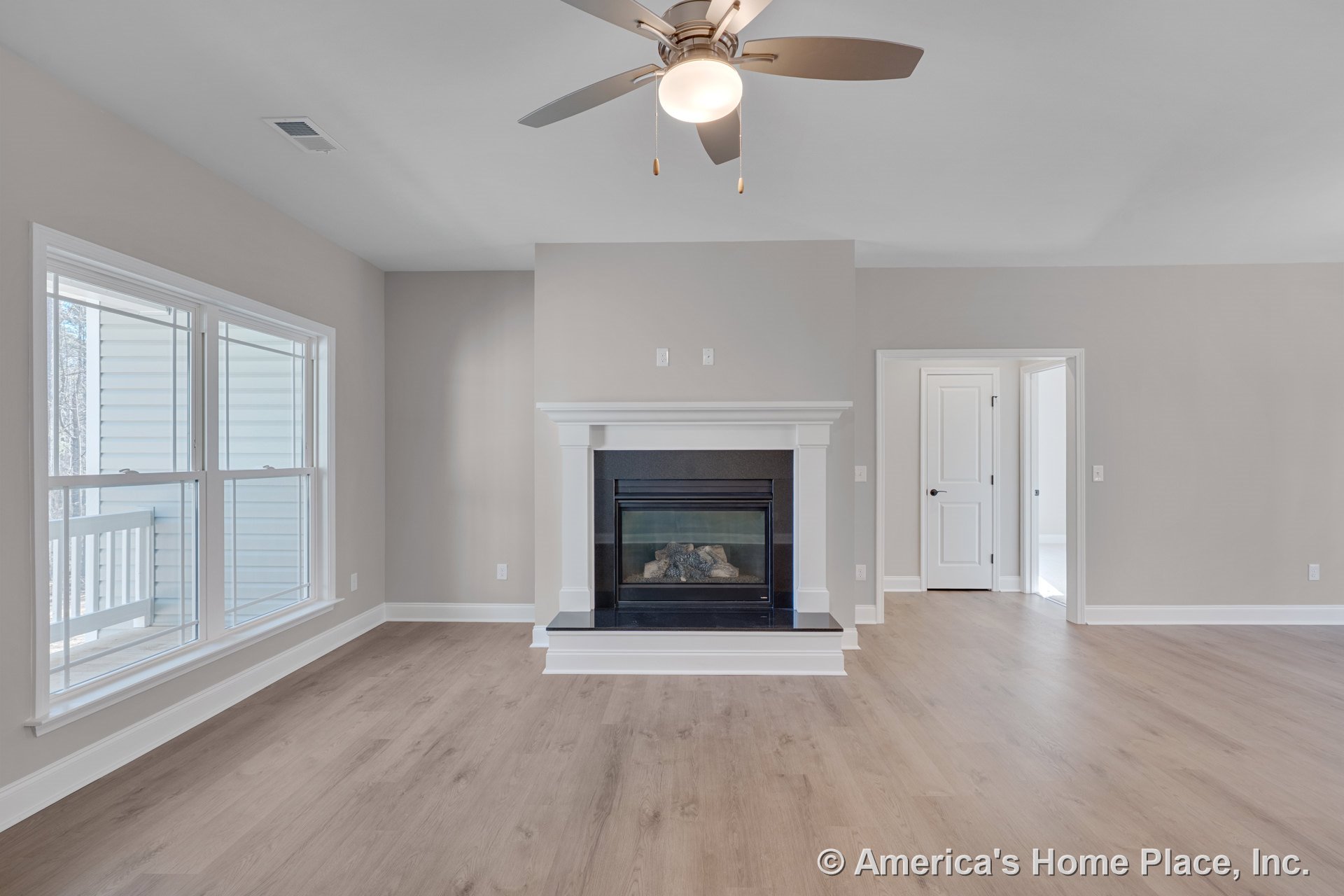 Central gas fireplace with black surround and white trim, light wood flooring, large windows with white casing, ceiling fan with built-in light, neutral wall paint, white