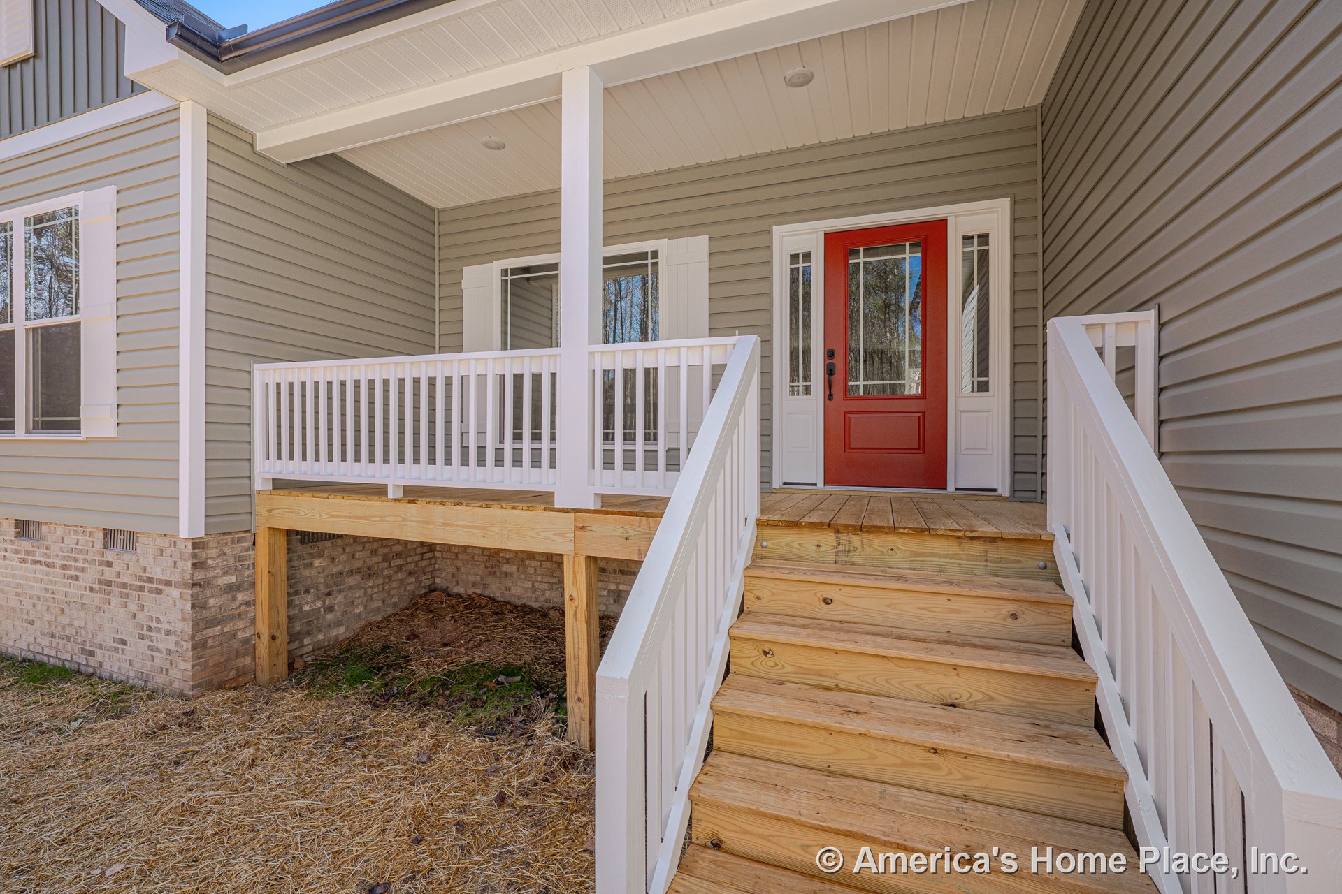 Covered front porch with wooden steps, white railings, red entry door flanked by sidelights, brick foundation, gray vinyl siding, and outdoor lighting fixtures.