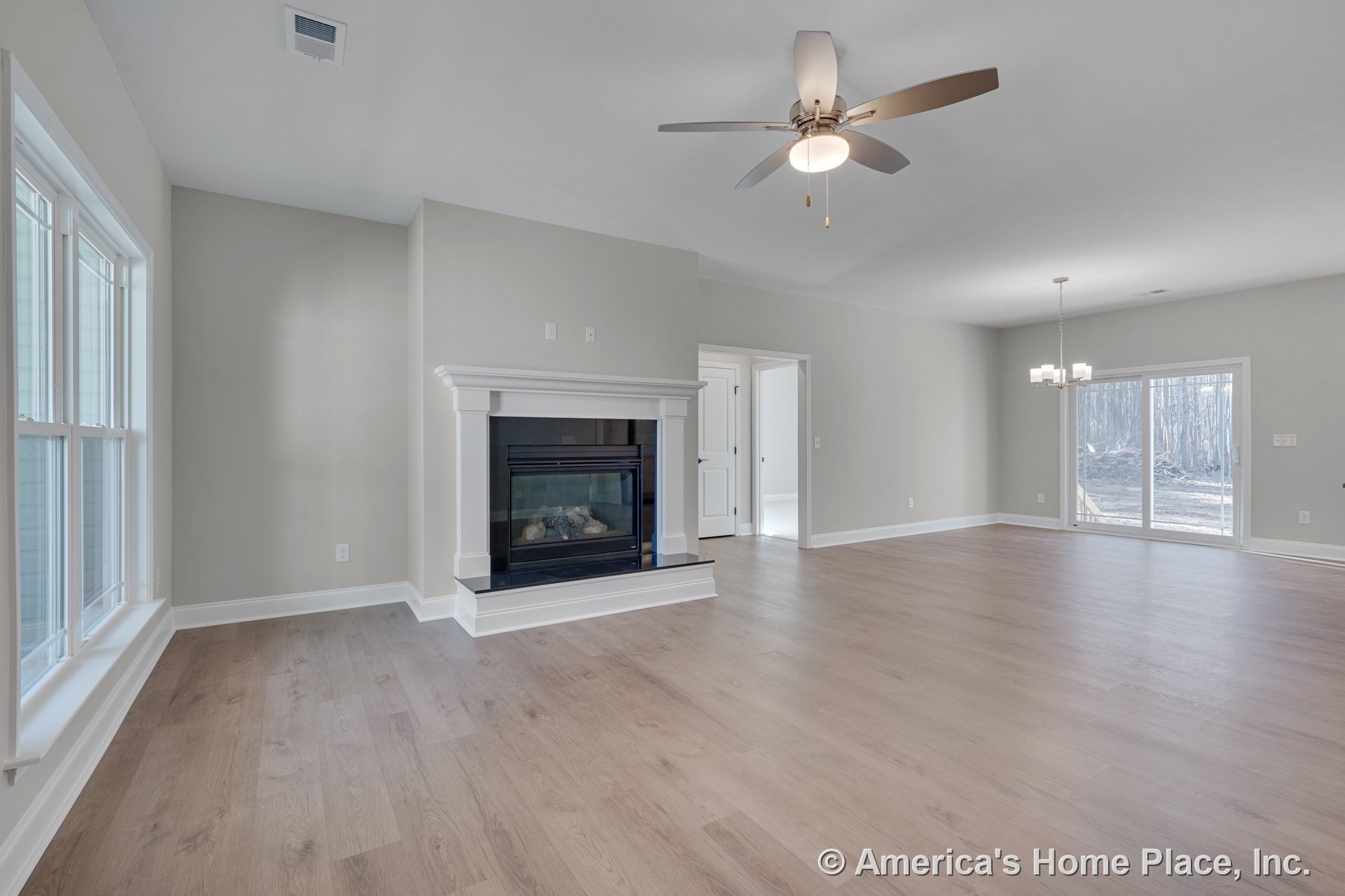 Gas fireplace with white mantel, light wood flooring, large double windows, ceiling fan with light, sliding glass door, modern chandelier, neutral walls, open layout.