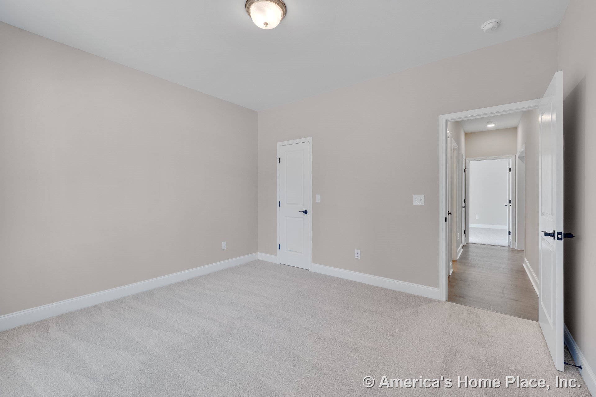 Bedroom with light carpet flooring, beige walls, white baseboards and trim, flush mount ceiling light, white paneled doors, and adjacent hallway featuring wood flooring.