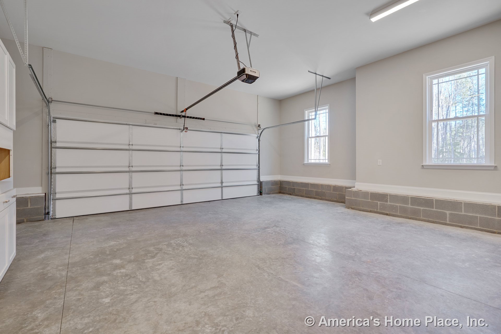 Spacious garage with sectional overhead door, concrete slab flooring, two double-hung windows, block foundation wall, ceiling-mounted garage door opener, and built-in cabinetry