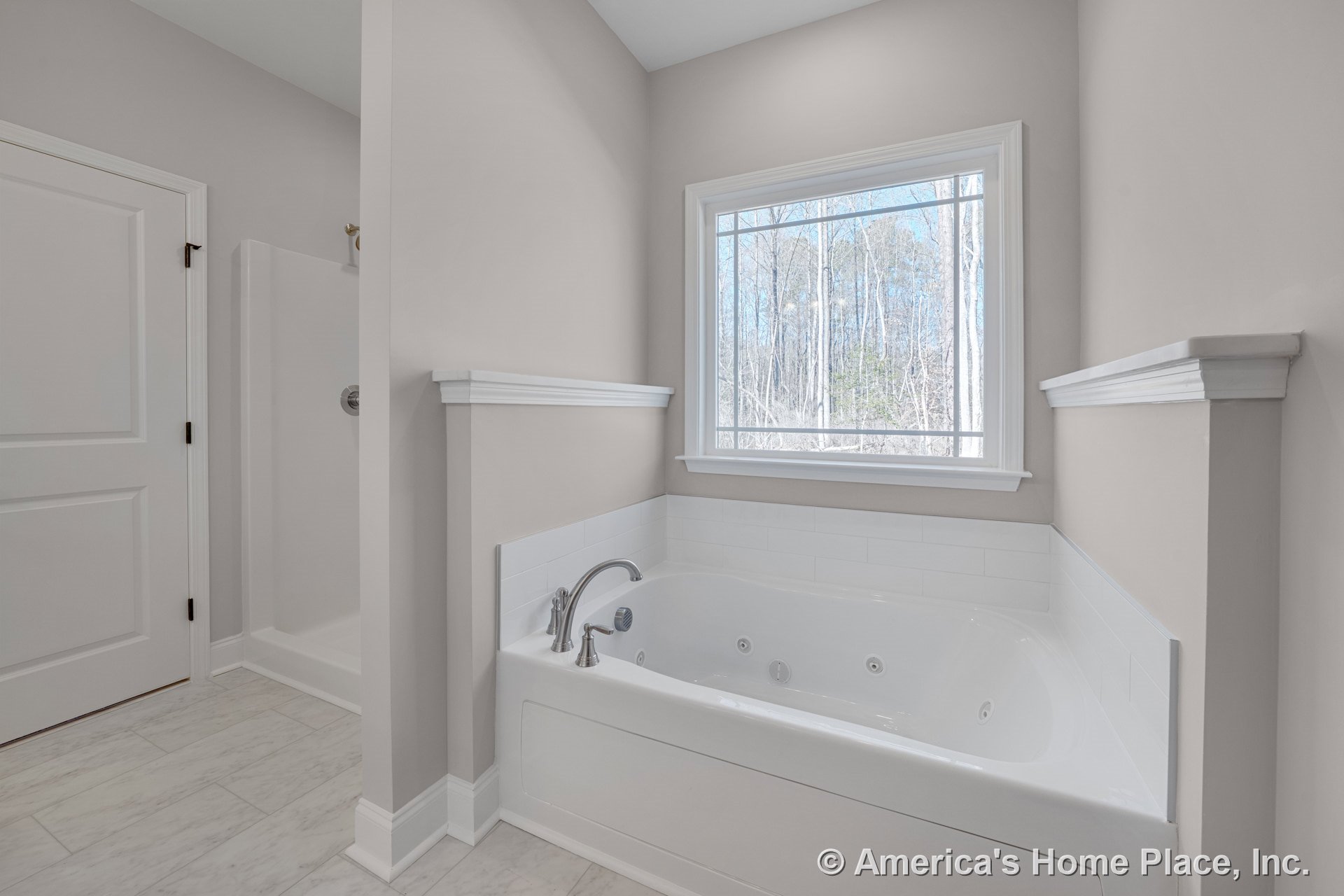 Jetted bathtub with tile surround positioned beneath a large window with trim, separate glass-enclosed shower stall, marble-look tile flooring, white painted door, crown molding