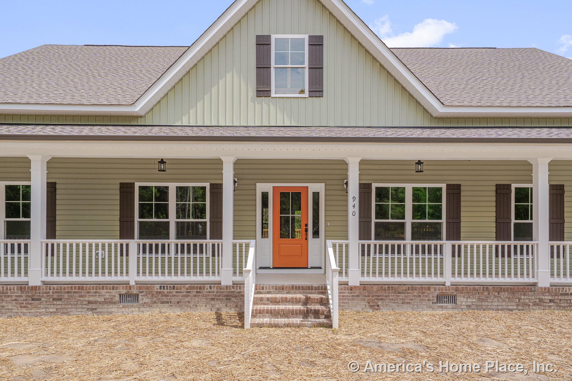 Covered front porch with white railings and columns, brick steps leading to an orange entry door, double-hung windows with dark shutters, exterior lighting fixtures, light siding