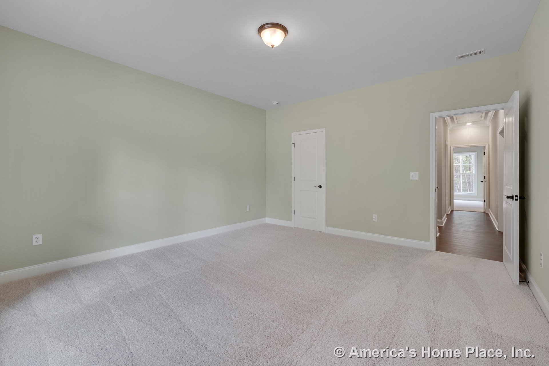 Bedroom with light carpet flooring, pale green walls, white trim and baseboards, flush mount ceiling light, white paneled doors, and adjacent hallway with wood flooring.